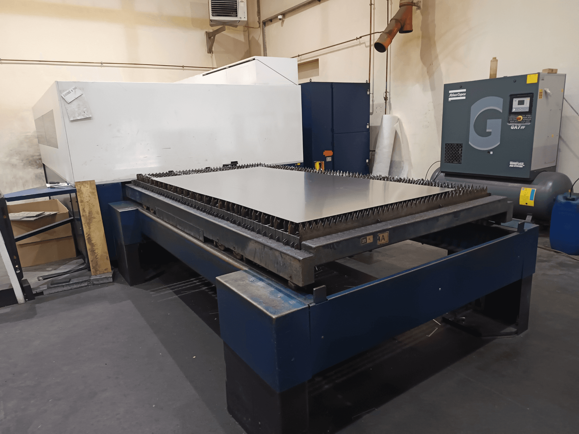 Máquina de corte industrial com mesa de metal, vista frontal. Apresenta arestas vivas, compressor de ar à direita e estrutura em aço.