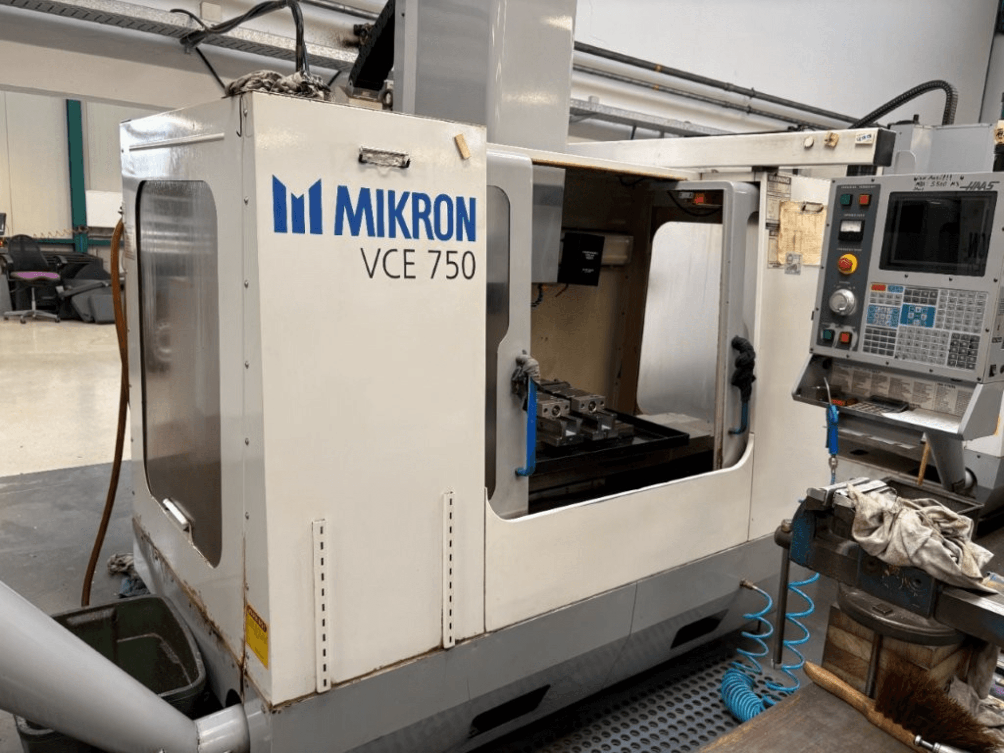Vista frontal da fresadora CNC Mikron VCE 750, com uma mesa de trabalho maquinada e um painel de controlo com vários botões e ecrãs.