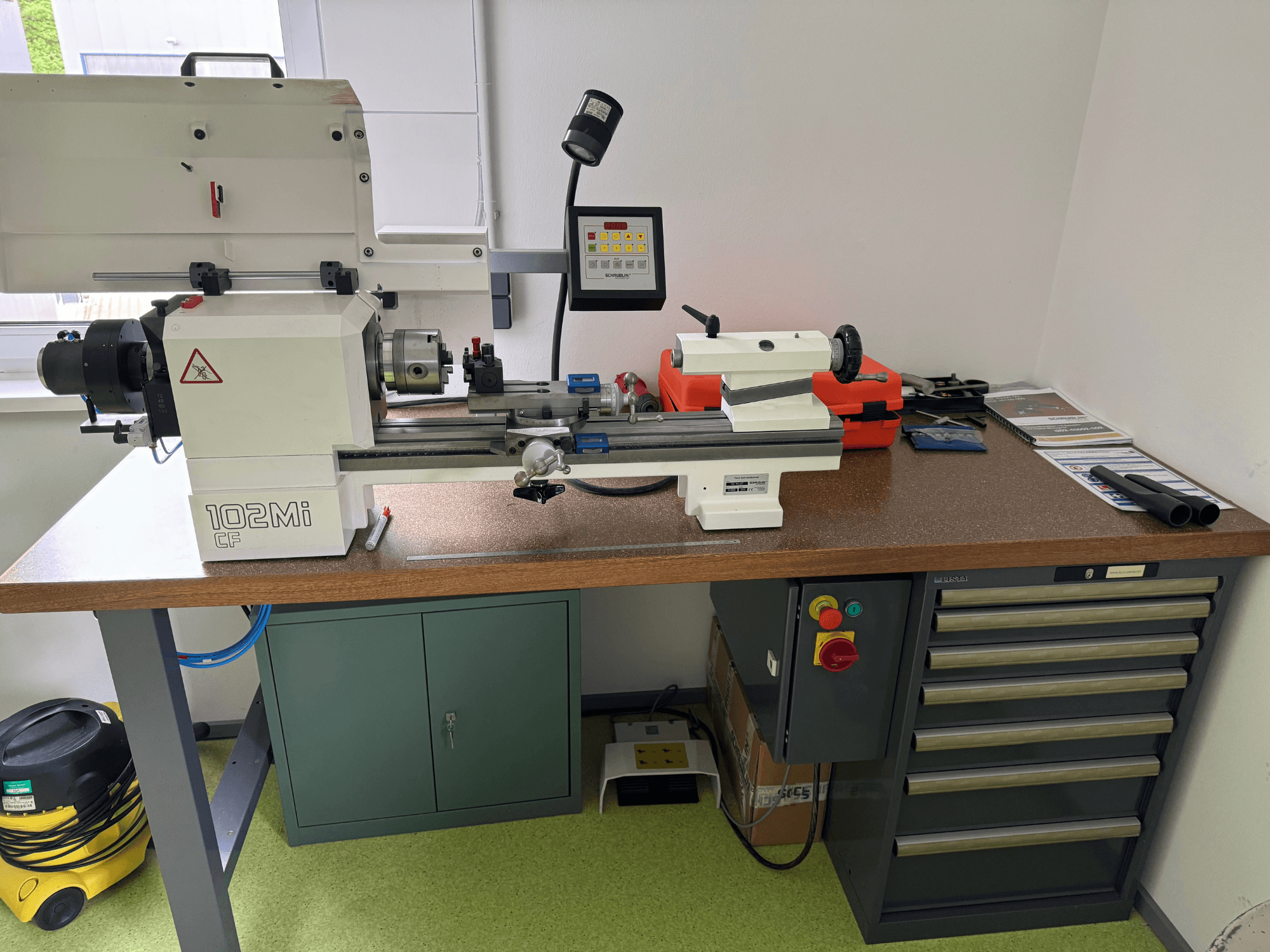 Torno horizontal SCHAUBLIN 102Mi W25 numa oficina, vista frontal com detalhes do painel de controlo e da bancada de trabalho.
