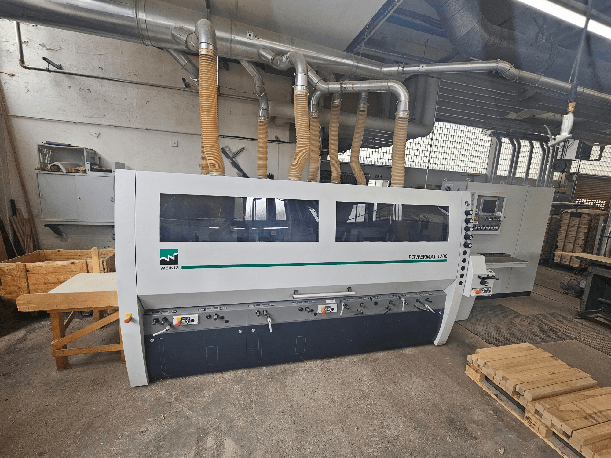 Máquina para trabalhar madeira Weinig Powermat 1200, vista frontal, com painel de controlo e pilhas de madeira numa oficina.