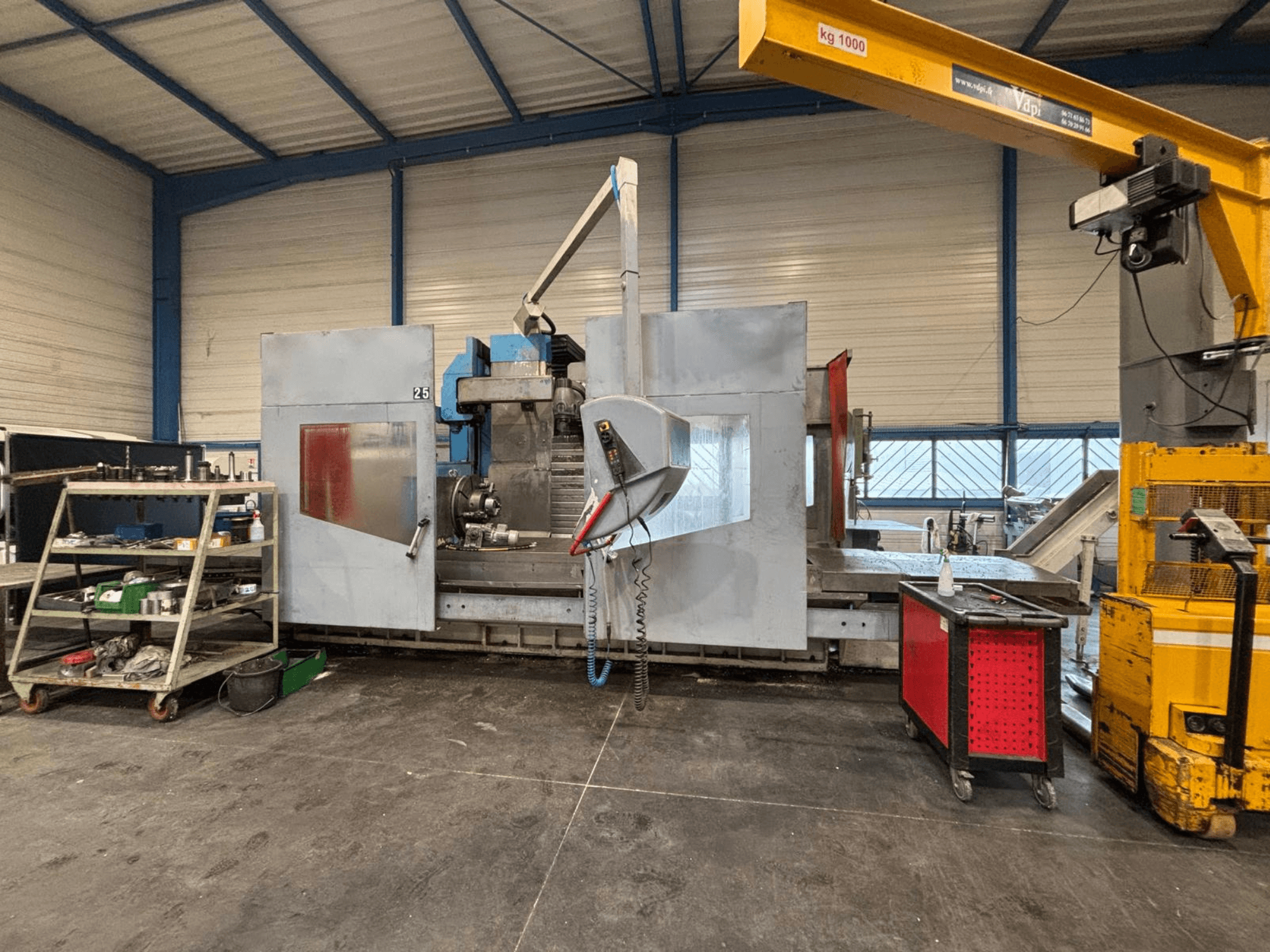 Fresadora CNC vista de lado numa oficina; ferramentas visíveis e prateleiras de arrumação. Marca e modelo não especificados.