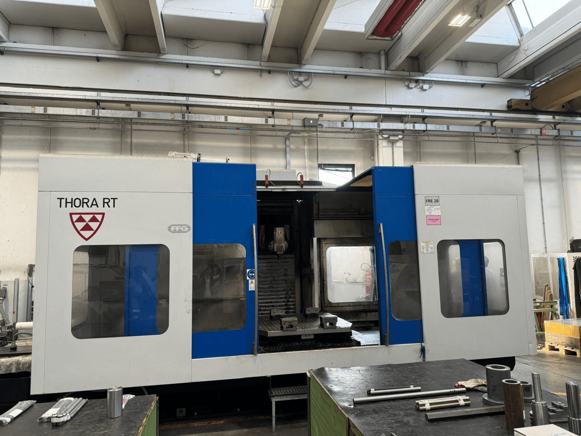 Máquina CNC THORA RT vista de frente, com um exterior azul e branco, janelas transparentes e ferramentas montadas.