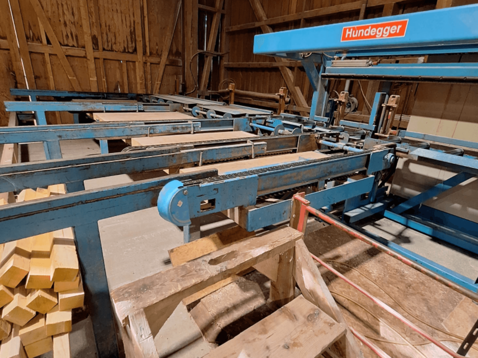 Centro de usinagem de madeira CNC HUNDEGGER K2-A num celeiro, com estrutura metálica azul, sistema de transporte e vigas de madeira empilhadas.