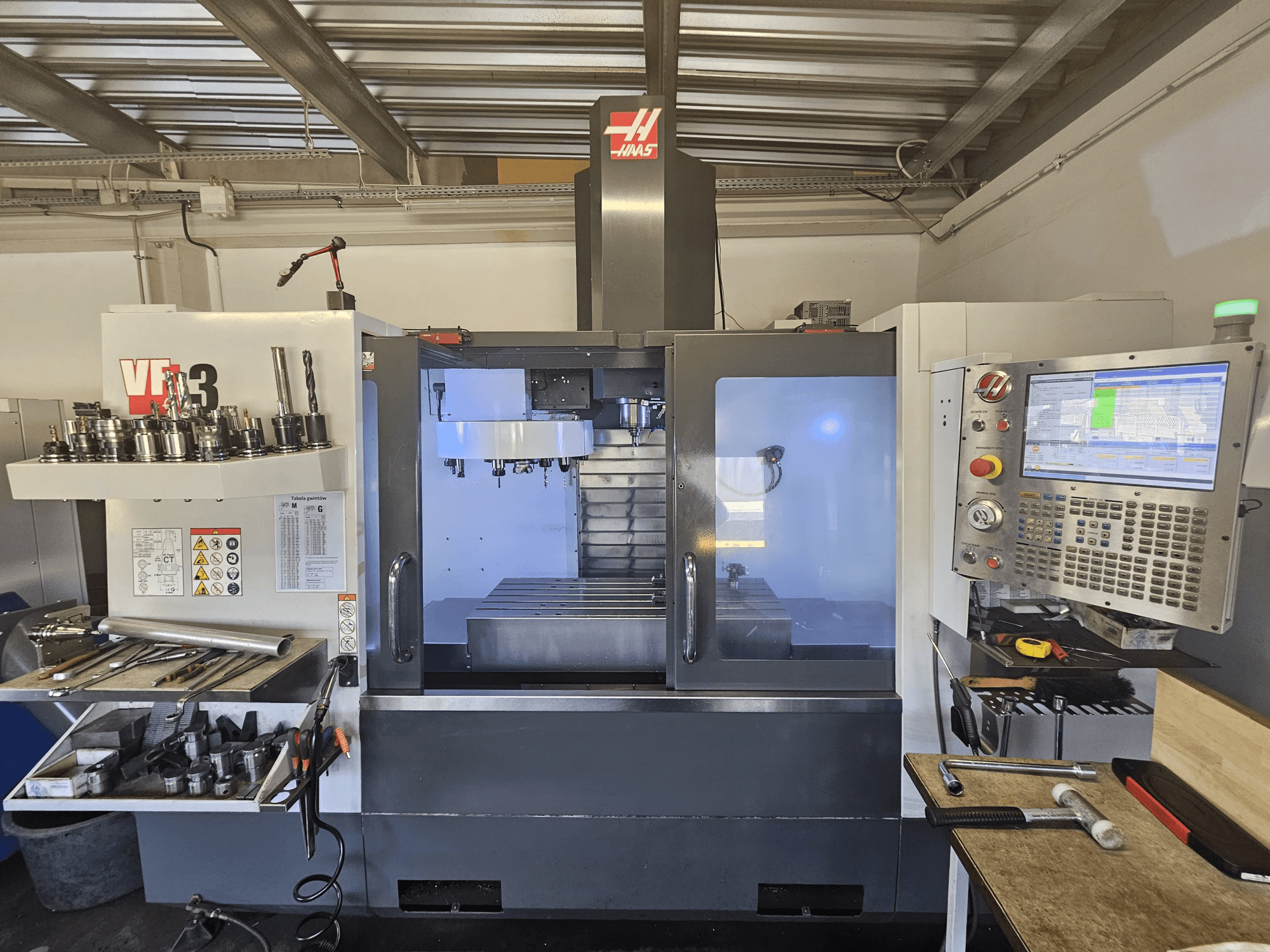 Máquina CNC Haas VF-3 numa oficina, vista frontal mostrando ferramentas, controlos e área de fresagem com interior iluminado.