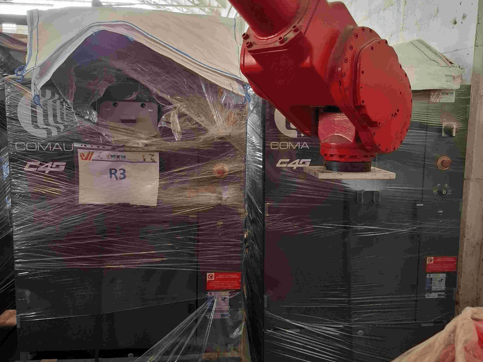 Braço robótico Comau C4G visto de frente, embrulhado em plástico, com a etiqueta "R3" e autocolantes de precaução visíveis na máquina.