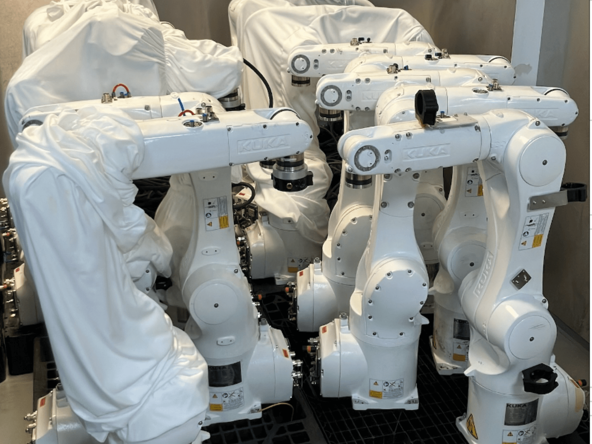 Braços robóticos KUKA KR 10 R900-2 cobertos com tecido branco, vistos de um ângulo superior, mostrando várias unidades em armazenamento.