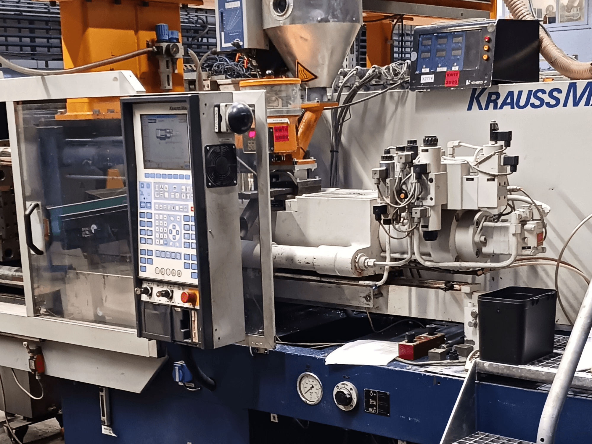 Máquina de moldagem por injeção KraussMaffei com painel de controlo, unidades hidráulicas e ferramentas em primeiro plano, vista frontal.