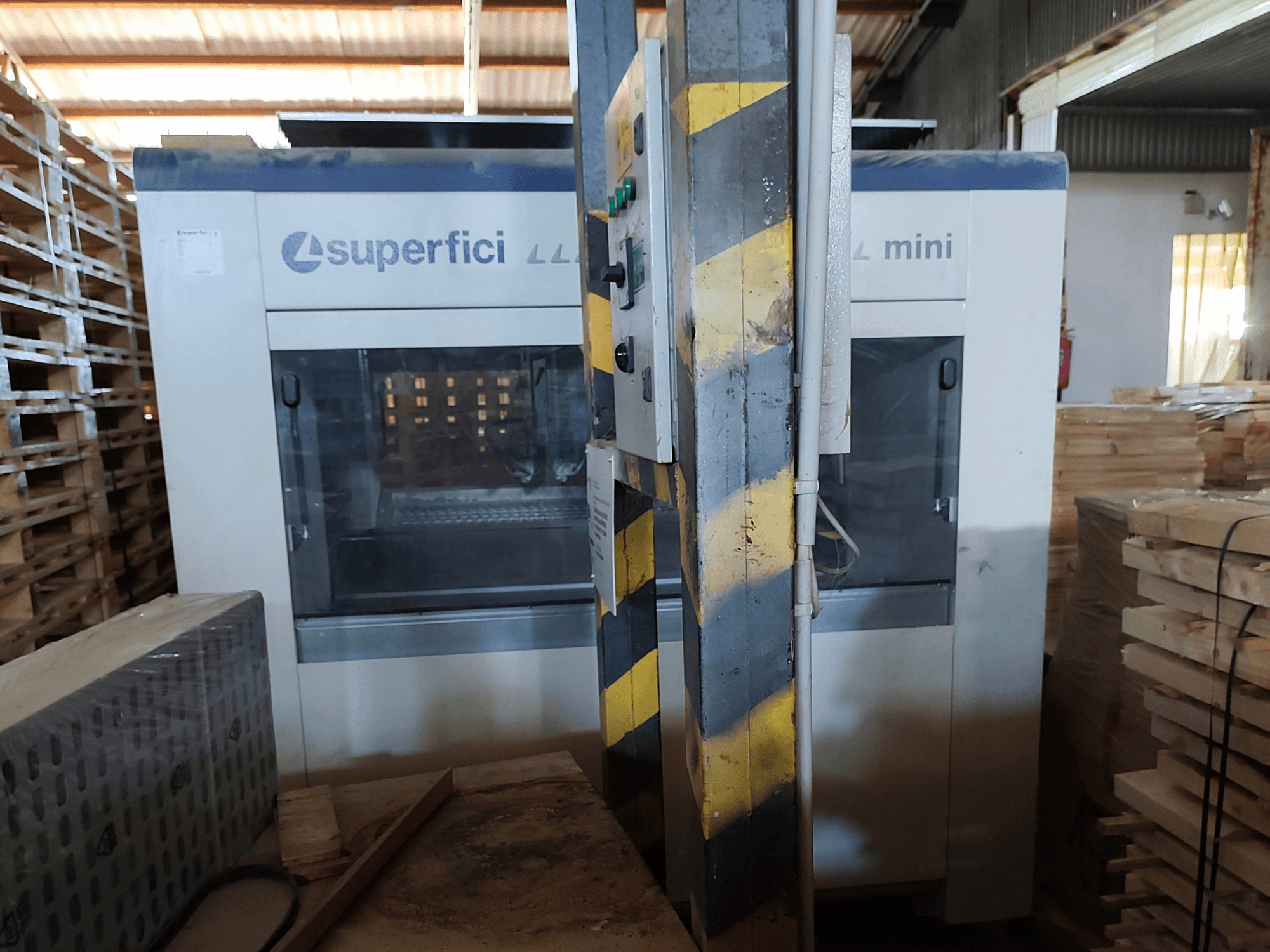 Máquina de acabamento de madeira SCM Superfici Mini vista de frente, mostrando o painel de controlo e as paletes de madeira à sua volta.