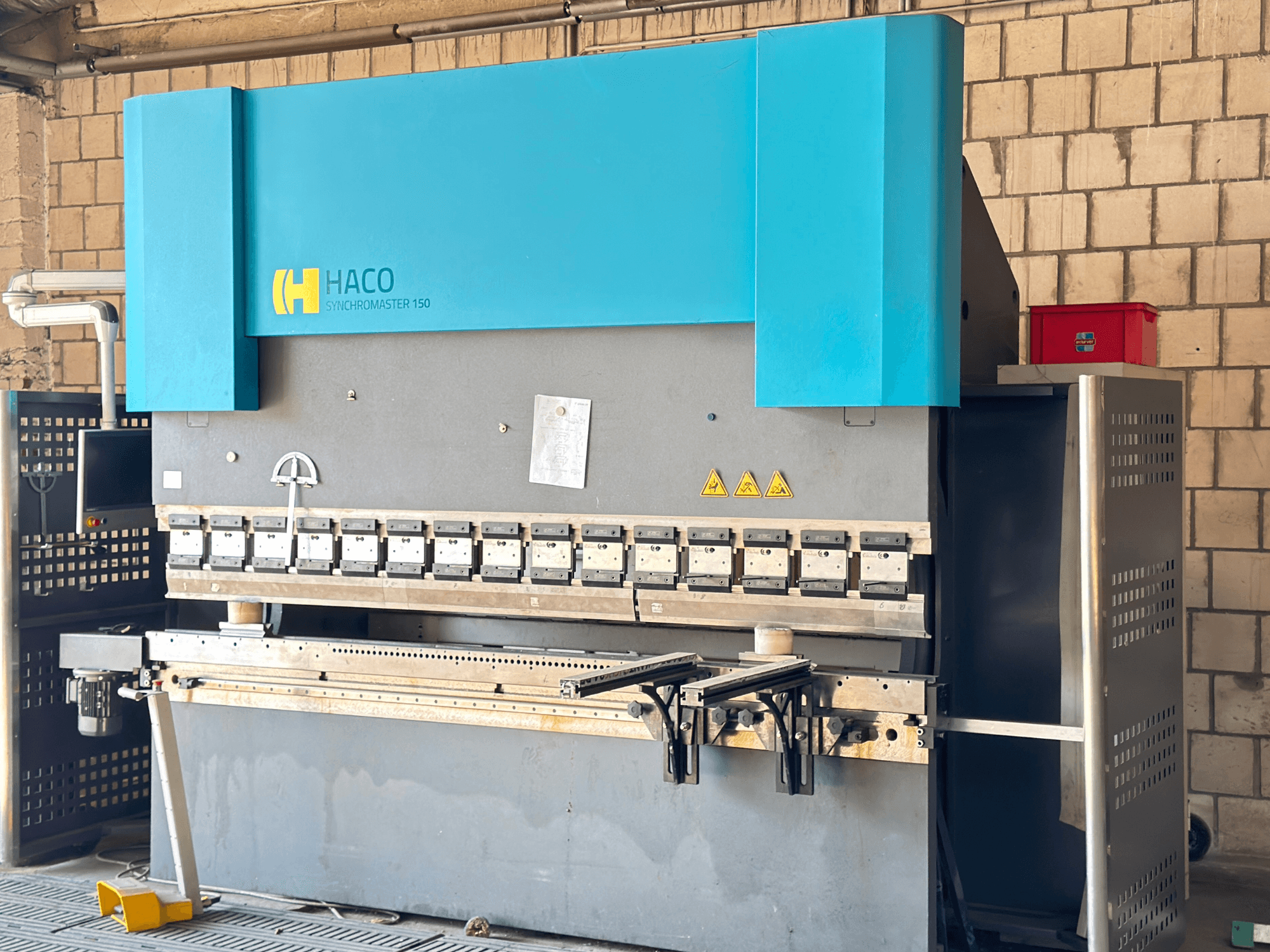 Prensa dobradeira HACO SRM30150, vista frontal; inclui painel de controlo e matrizes de fixação, com etiquetas de aviso de segurança visíveis.