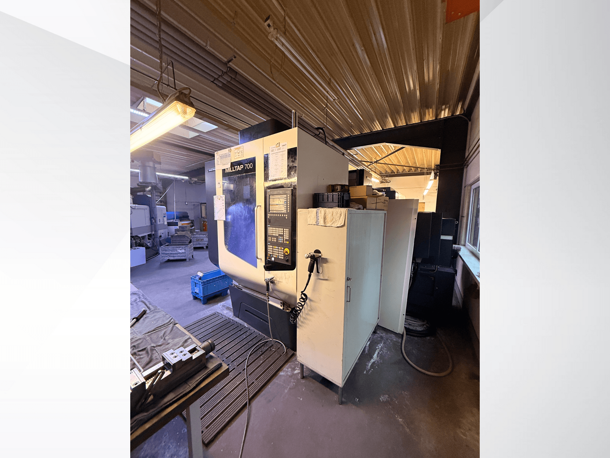 Máquina CNC Milltap 700 na oficina; a vista frontal mostra o painel de controlo, as prateleiras de ferramentas e as áreas de armazenamento com iluminação acima.