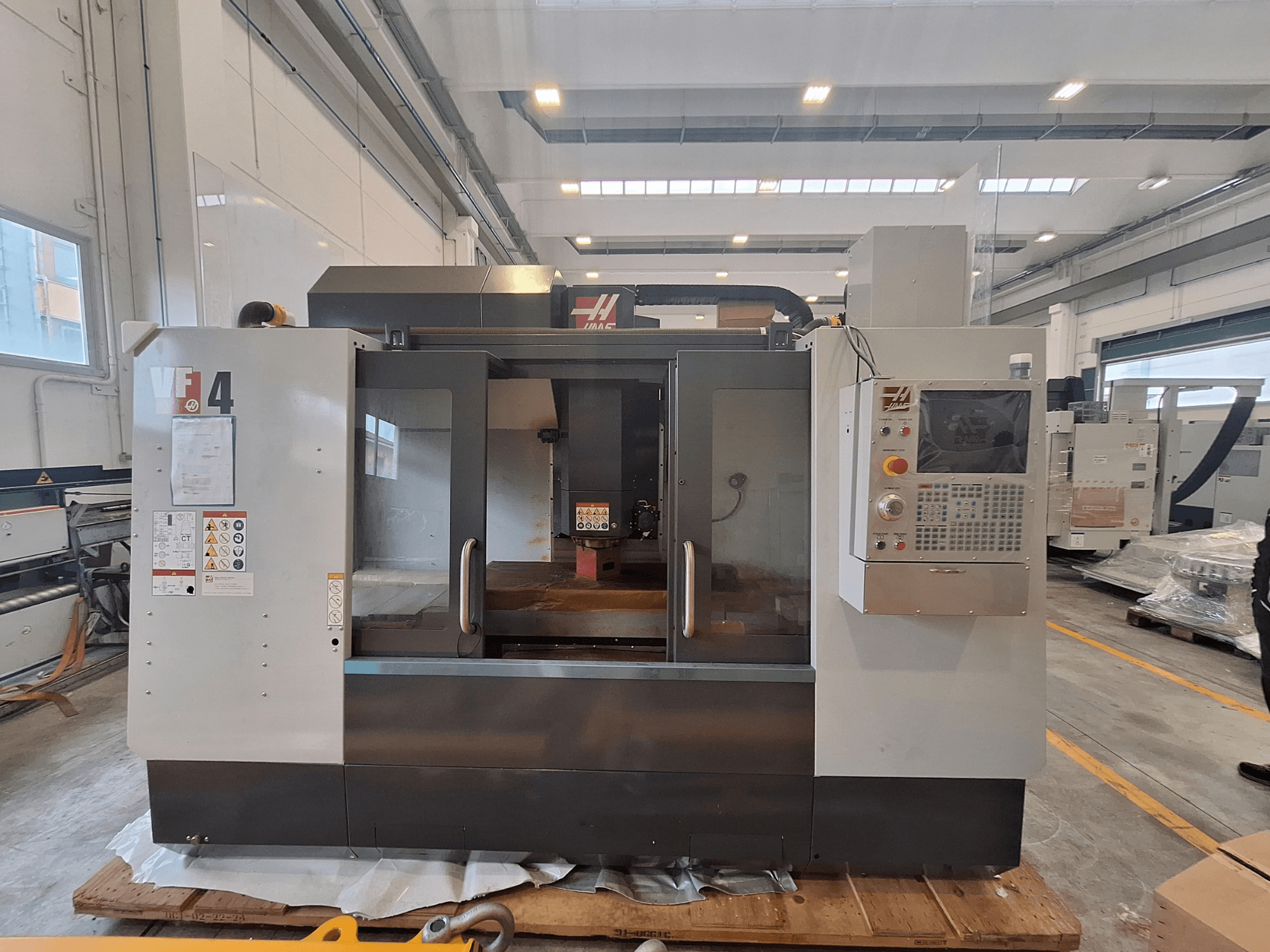 Centro de maquinagem vertical HAAS VF-4, vista frontal, mostrando o painel de controlo, a área de trabalho e as etiquetas de segurança num ambiente de armazém.