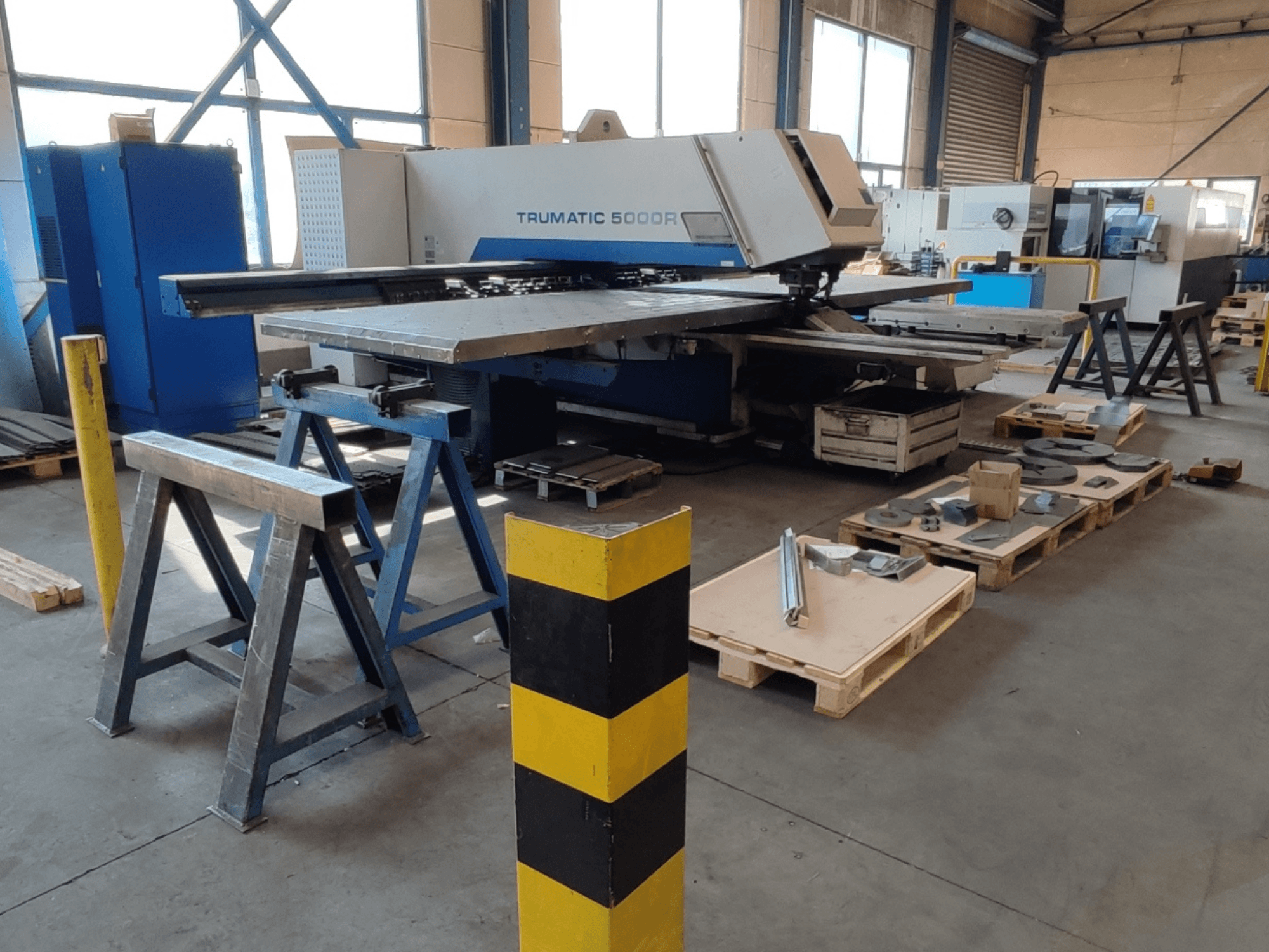 Máquina de perfuração CNC TRUMPF Trumatic 5000 R numa oficina, vista lateral com ferramentas e materiais em paletes nas proximidades.