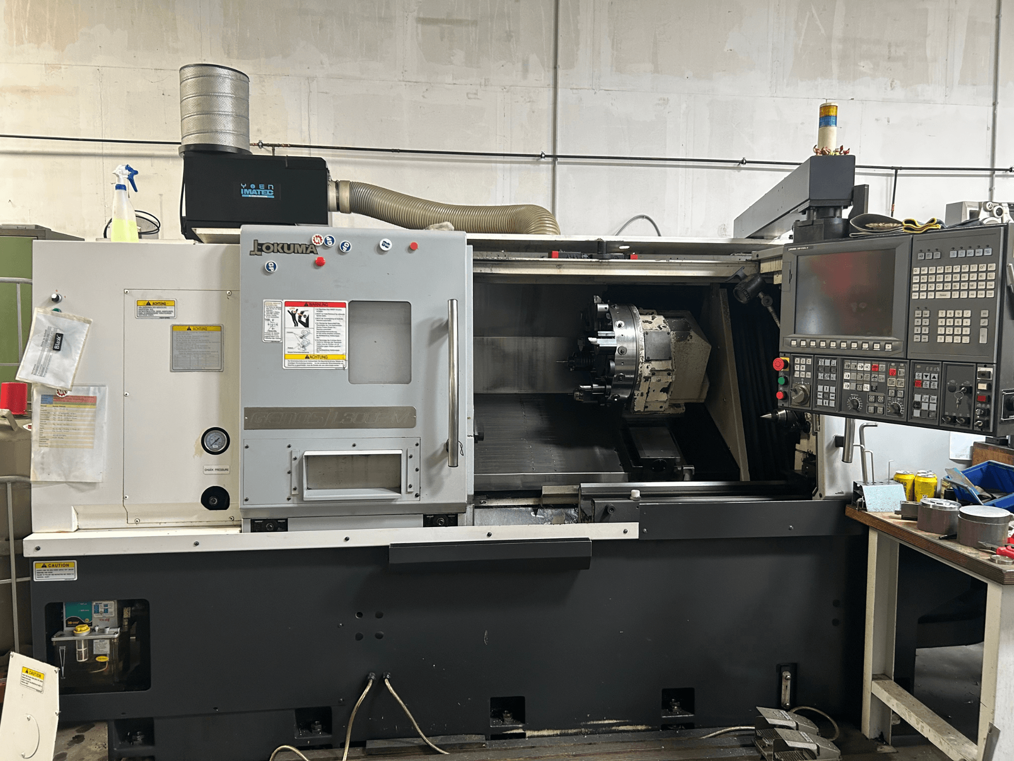 Vista frontal de um torneiro horizontal Okuma Genos L300E-M com painel de controlo, fuso visível e várias ferramentas de configuração.