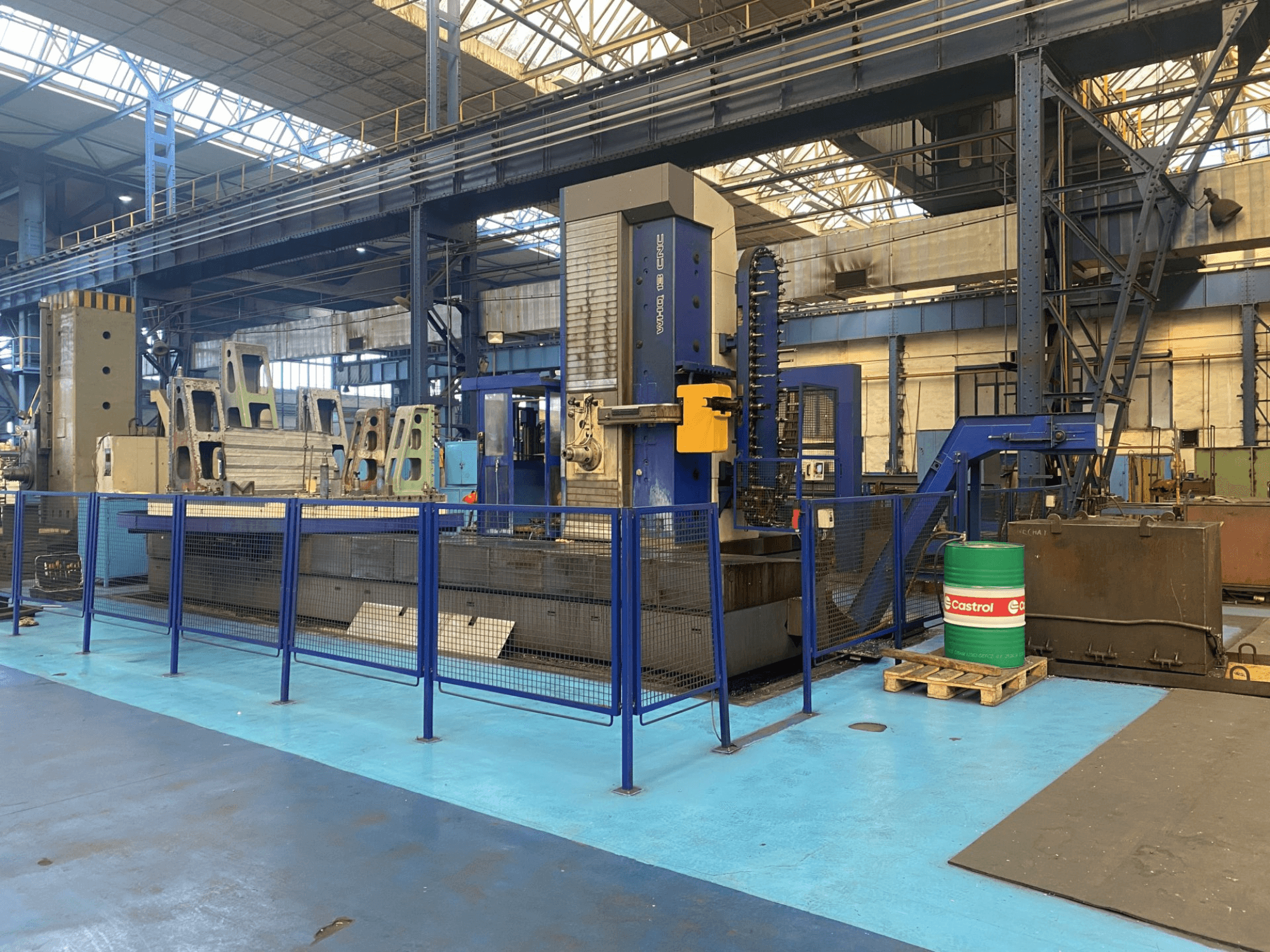 Máquina de furar CNC TOS WHQ 13 numa oficina, vista frontal, mostrando a estrutura azul e a vedação de segurança circundante.