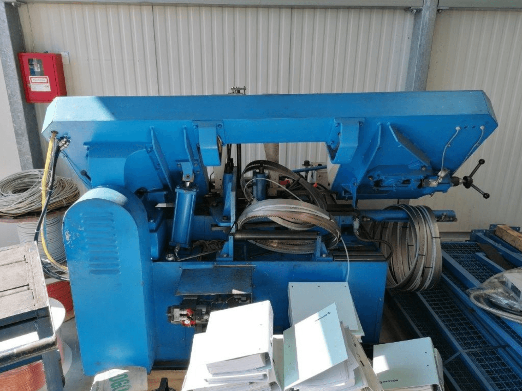 Máquina industrial azul com componentes hidráulicos, vista lateral, inclui mangueiras e folhas de metal numa superfície.