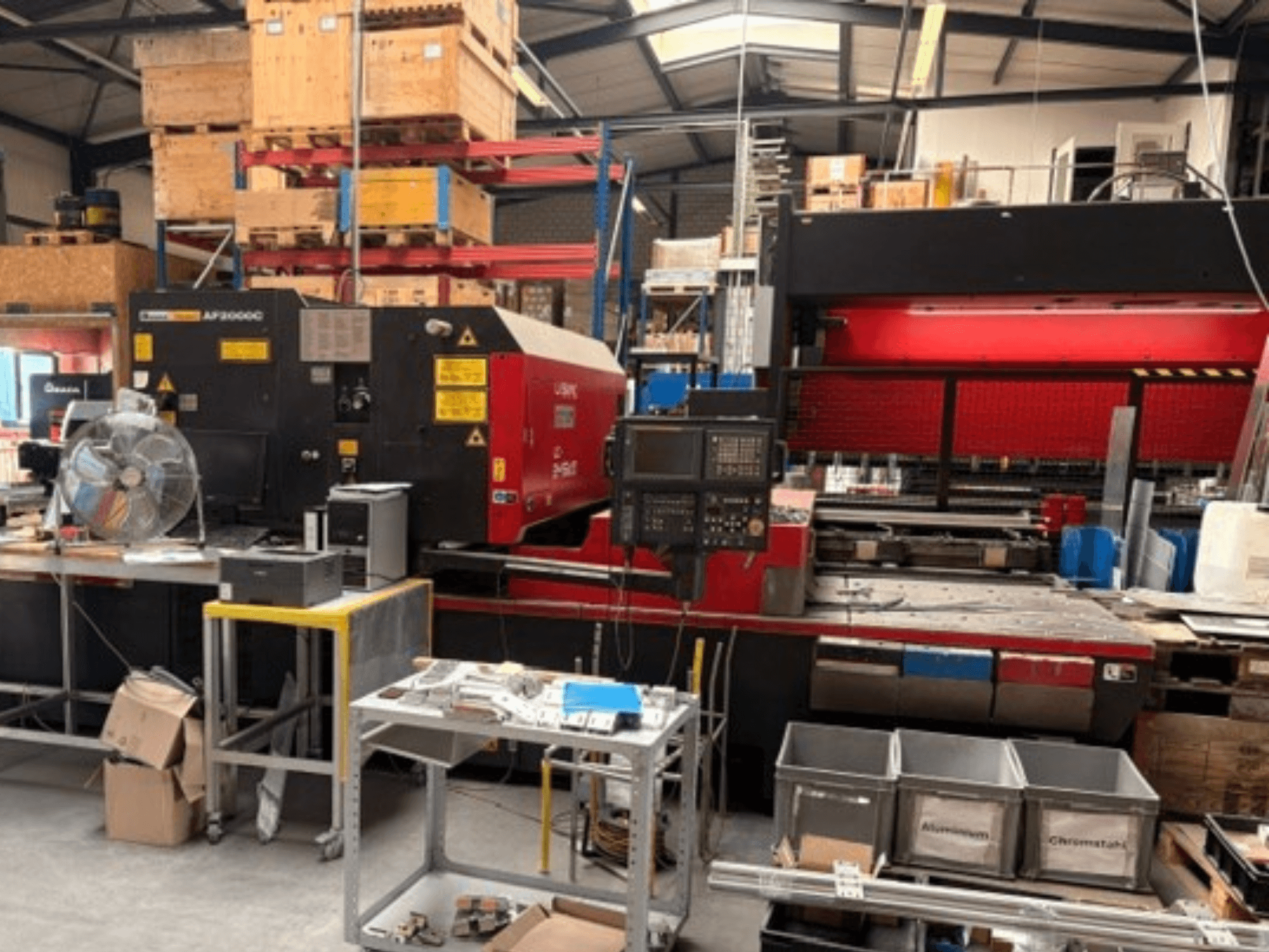 Máquina de corte a laser de CO₂ AMADA LC-2415αIII num ambiente de oficina, mostrando a vista frontal, o painel de controlo e o espaço operacional.