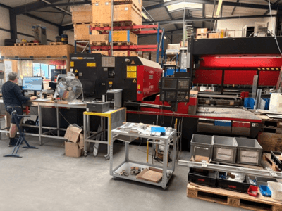 Máquina de corte a laser de CO₂ AMADA LC-2415αIII num ambiente de oficina, com uma estação de trabalho informática visível em primeiro plano.