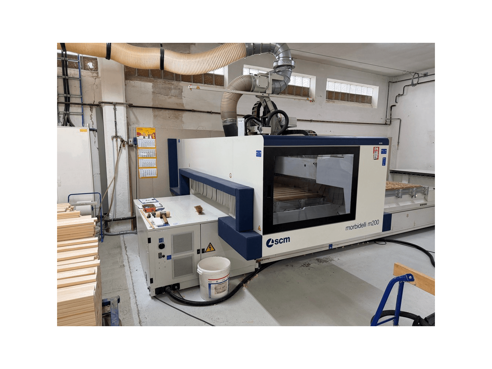 Centro de usinagem CNC SCM Morbidelli M200F numa oficina, vista lateral com peças de madeira na mesa e sistema de ventilação na parte superior.