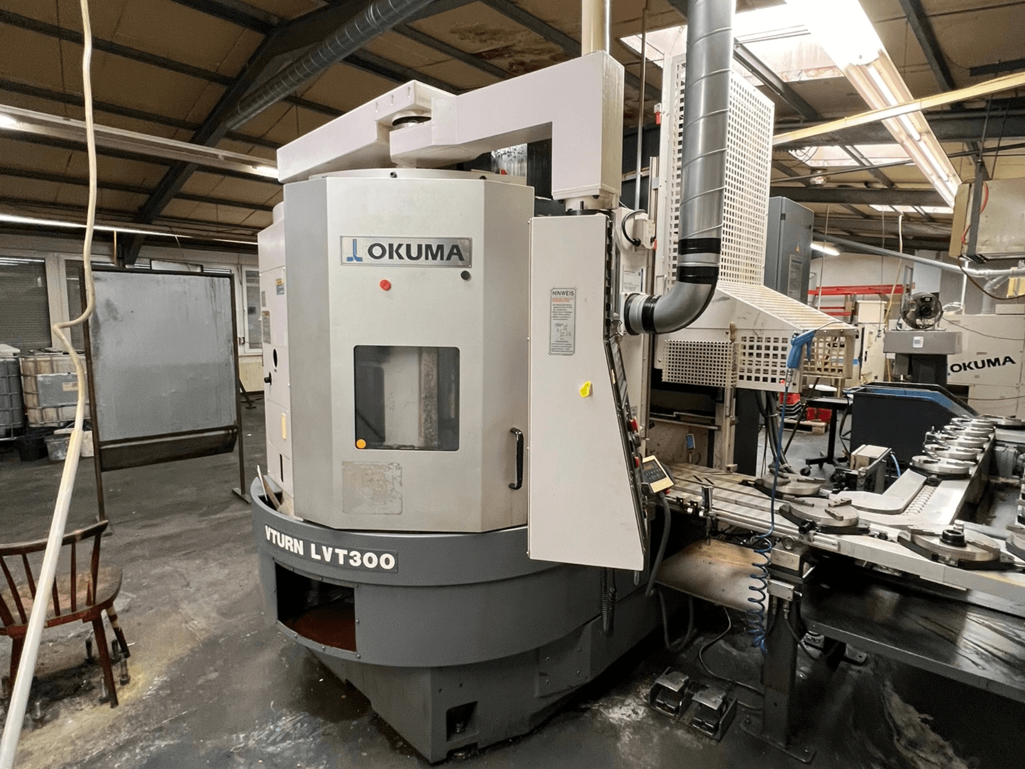 Torno CNC Okuma Vturn LVT300, vista lateral em ambiente industrial, mostrando o painel de controlo e o sistema de refrigeração.