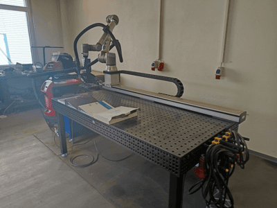 Célula de soldadura por impulsos FRONIUS TransSteel 4000 vista de lado, com um braço robótico, área de trabalho e ligações de alimentação.