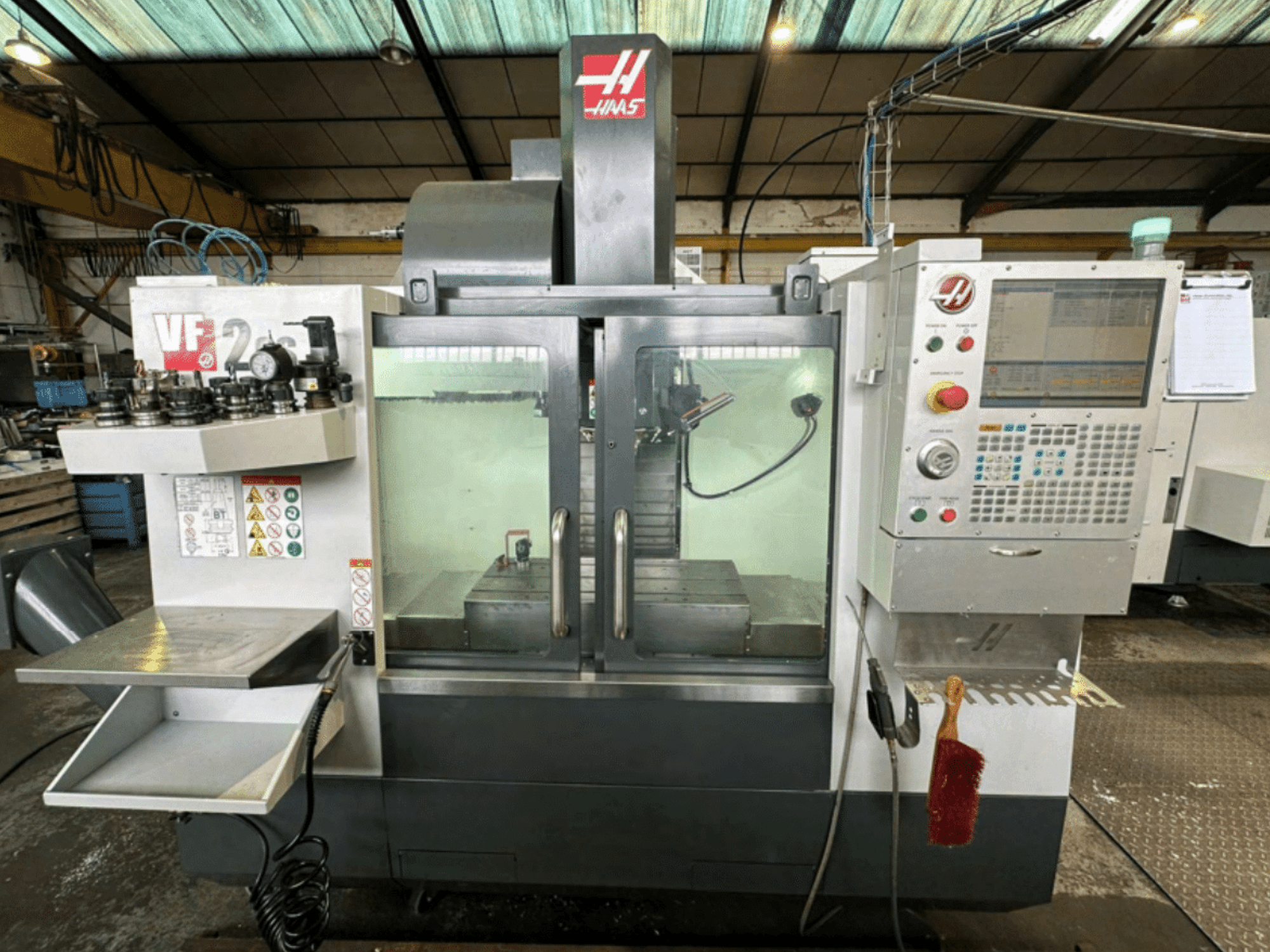 Vista frontal da máquina CNC Haas VF2, mostrando o painel de controlo, suportes de ferramentas e espaço de trabalho com etiquetas de segurança e uma escova.