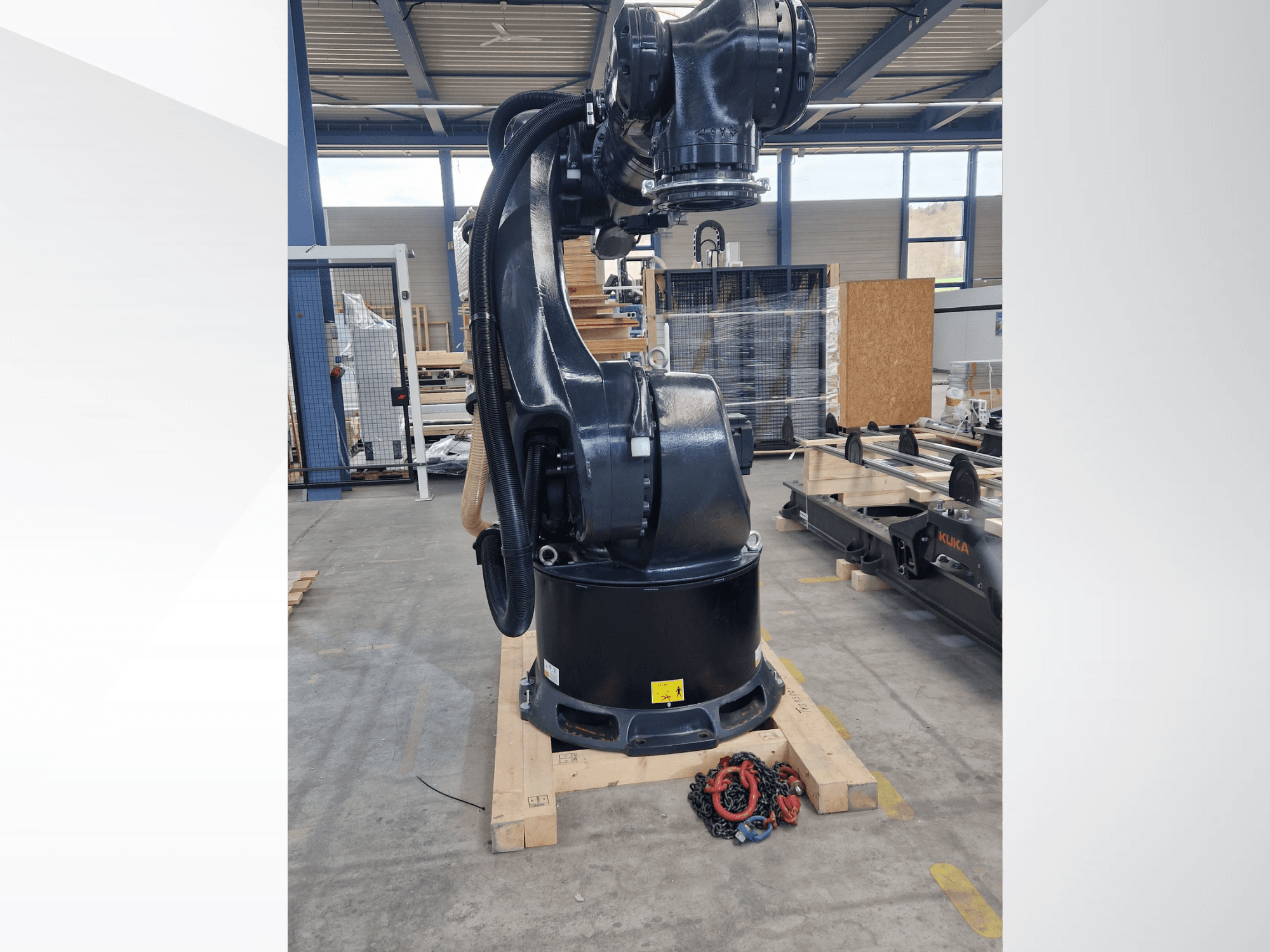 Braço robótico KUKA KR 340 R3330 num armazém, vista lateral que mostra a sua base e os cabos, com correntes em primeiro plano.