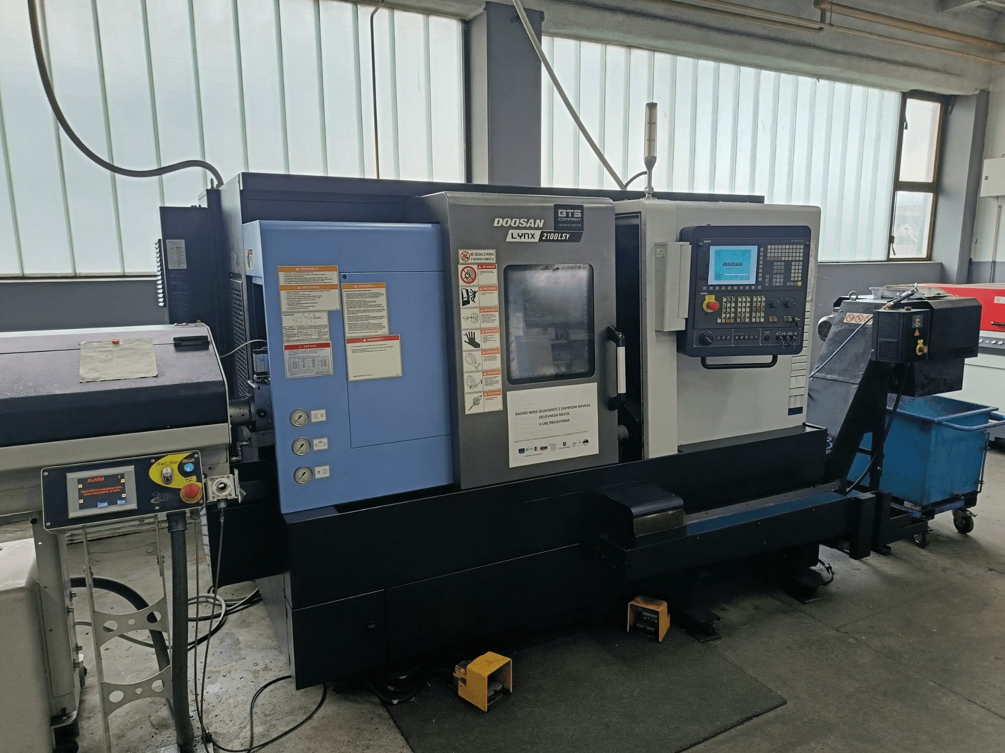 DOOSAN LYNX 2100 LSY Turn-Mill Centre, vista lateral mostrando o painel de controlo e os botões operacionais, com etiquetas informativas visíveis.