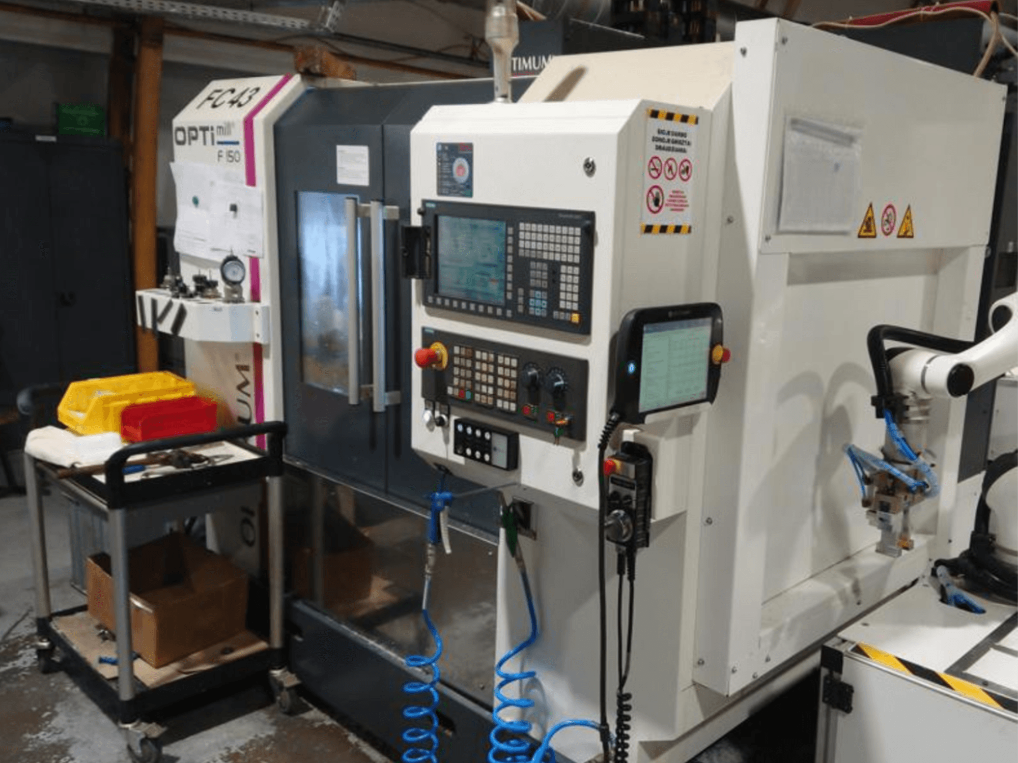 Painel de controlo da fresadora OPTIMUM F50 com mangueiras, ferramentas e um braço robótico visíveis numa oficina.