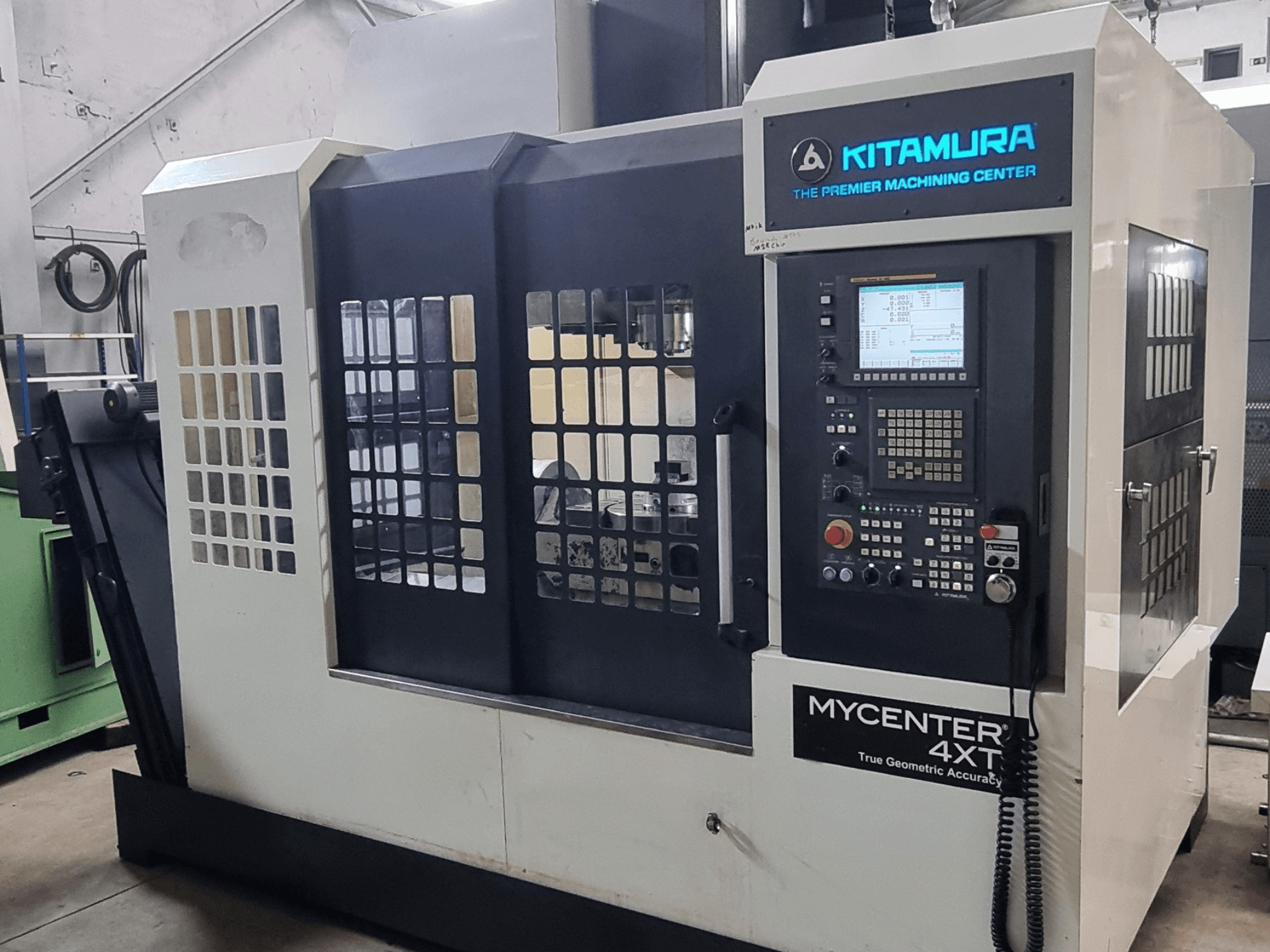 Texto alternativo: Centro de maquinagem vertical Kitamura MYCENTER 4XT, vista frontal, mostrando o painel de controlo e os detalhes da máquina num ambiente de oficina.