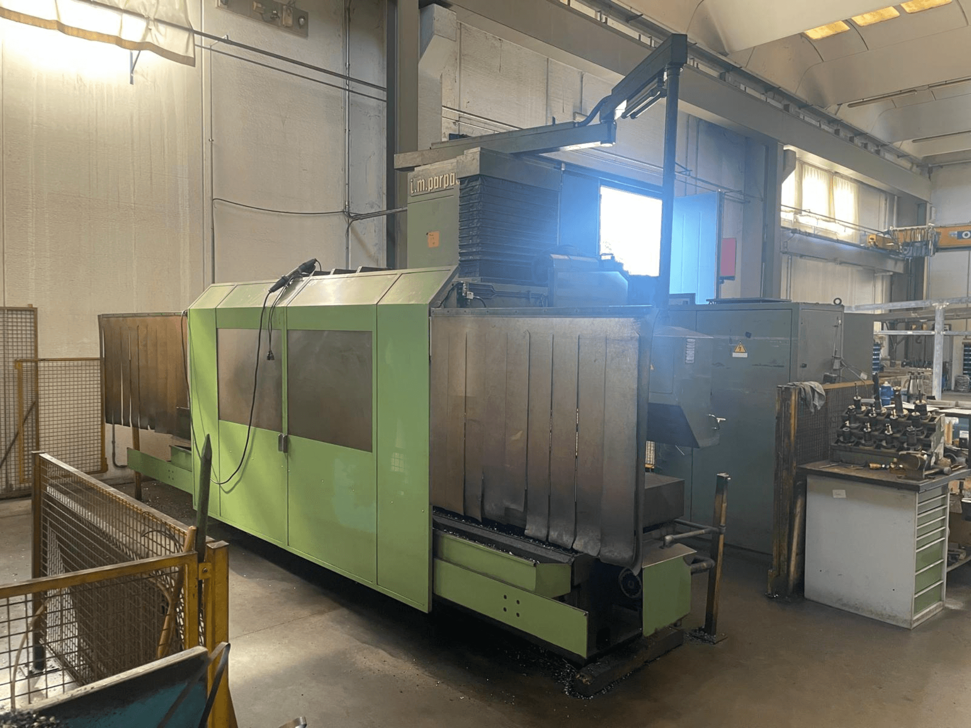 Máquina industrial verde com componentes metálicos visíveis, vista frontal; possui painel de controlo, avisos de segurança e posto de trabalho próximo.