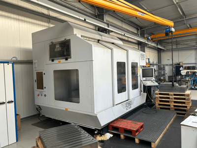 Máquina CNC Spinner VC 1650, vista frontal, com painel de controlo, janelas grandes e paletes empilhadas nas proximidades, numa oficina.