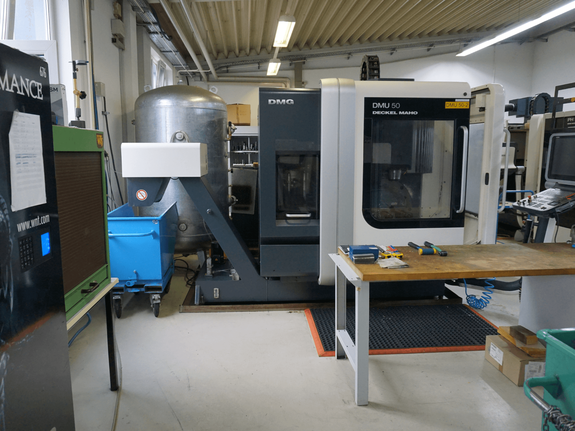 Fresadora DMG DMU 50 numa oficina, vista de frente, com ferramentas numa mesa de madeira e um caixote do lixo azul por perto.