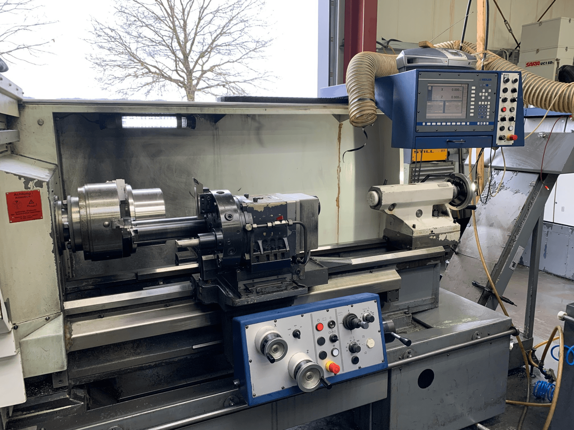 Torneiro horizontal Weiler E55X1000 numa oficina, mostrando o painel de controlo, o torno e o suporte de ferramentas. Vista frontal.