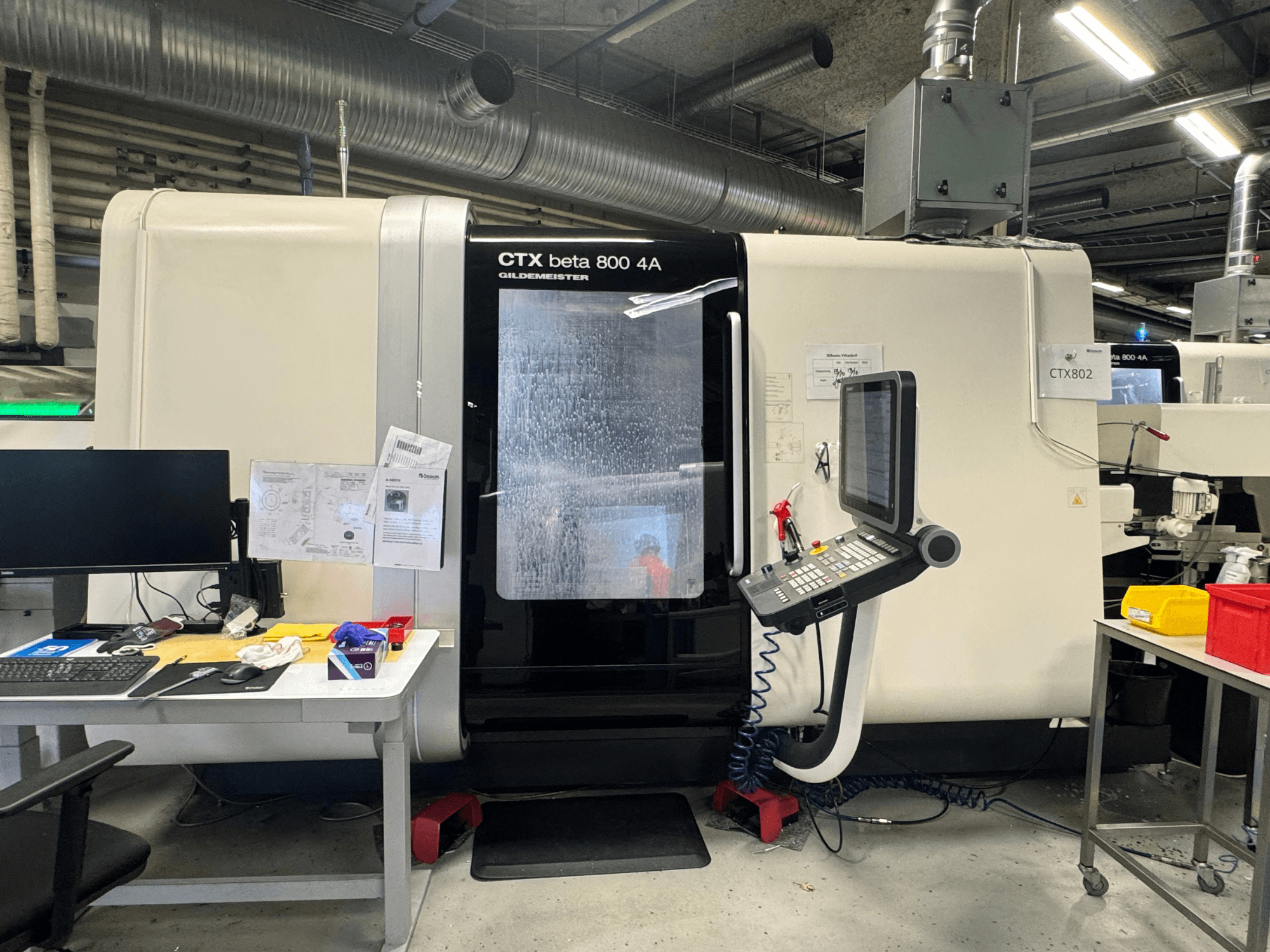 Máquina CNC CTX beta 800 4A numa oficina, vista lateral mostrando o painel de controlo e o espaço de trabalho cheio de ferramentas e documentos.