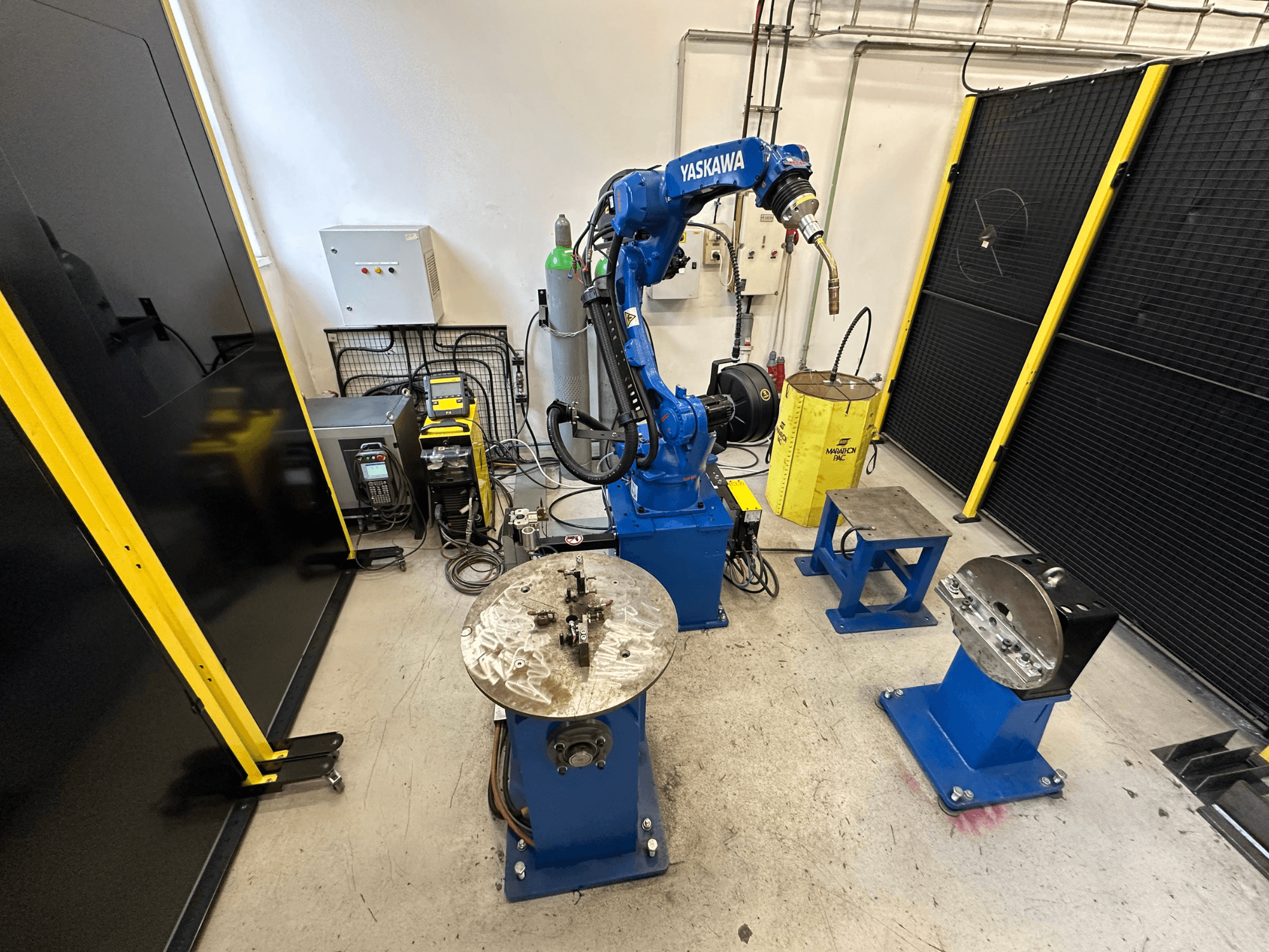 Braço robótico Yaskawa em azul com duas mesas de trabalho, equipamento de soldadura e barreiras de segurança; vista frontal de um espaço de trabalho industrial.