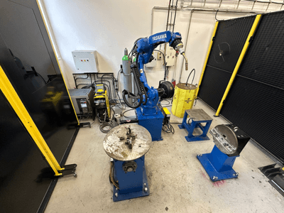 Braço robótico Yaskawa em azul com duas mesas de trabalho, equipamento de soldadura e barreiras de segurança; vista frontal de um espaço de trabalho industrial.