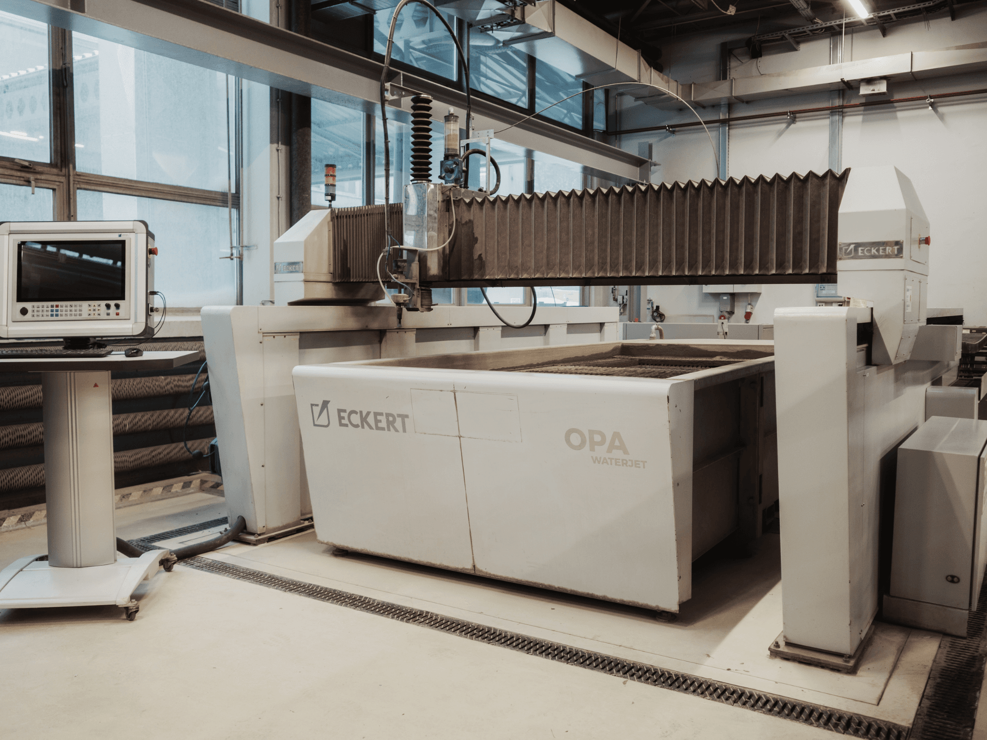 Máquina de corte por jato de água ECKERT OPAL numa oficina, vista de frente, com a consola de controlo nas proximidades.