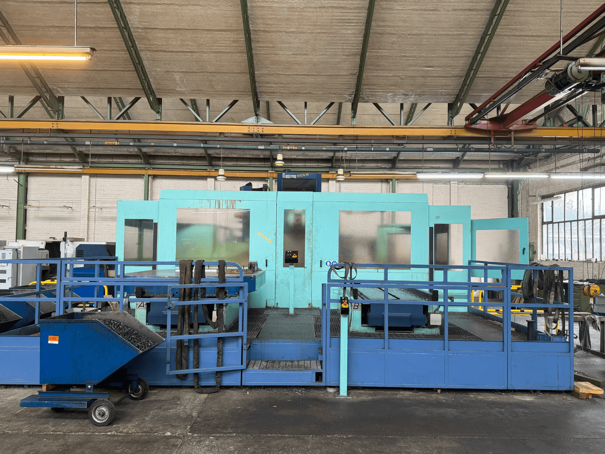 Máquina CNC industrial azul com painéis de proteção, vista frontal, com arrumação de ferramentas e um carrinho de recolha de resíduos na lateral.