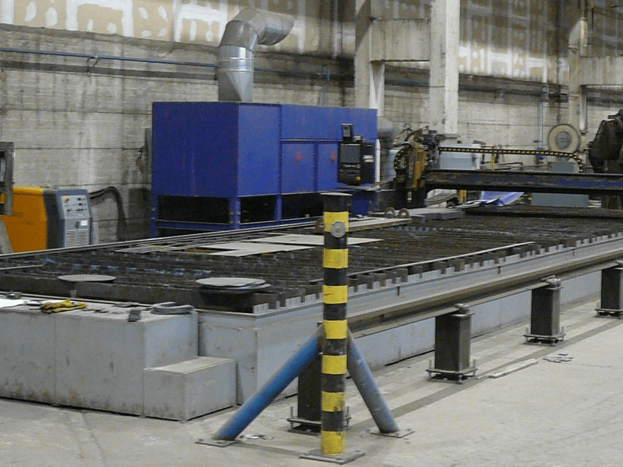 Configuração de máquina de corte de metal industrial, vista lateral, com uma caixa de controlo azul, faixas de segurança e superfície de trabalho em metal.