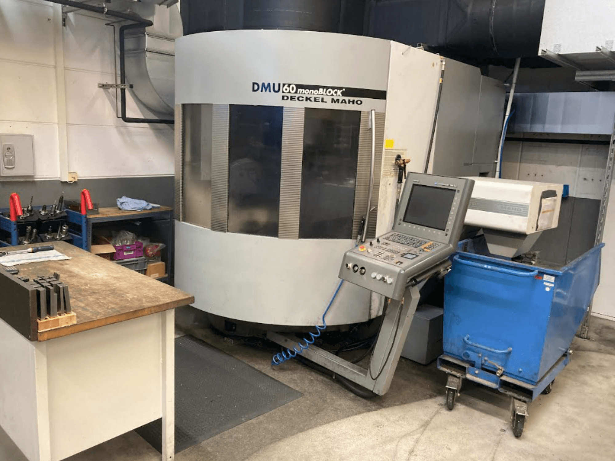 Centro de maquinação vertical DMG MORI DMU 60 Monoblock, vista frontal com painel de controlo, espaço de trabalho e carrinho de ferramentas azul visíveis.