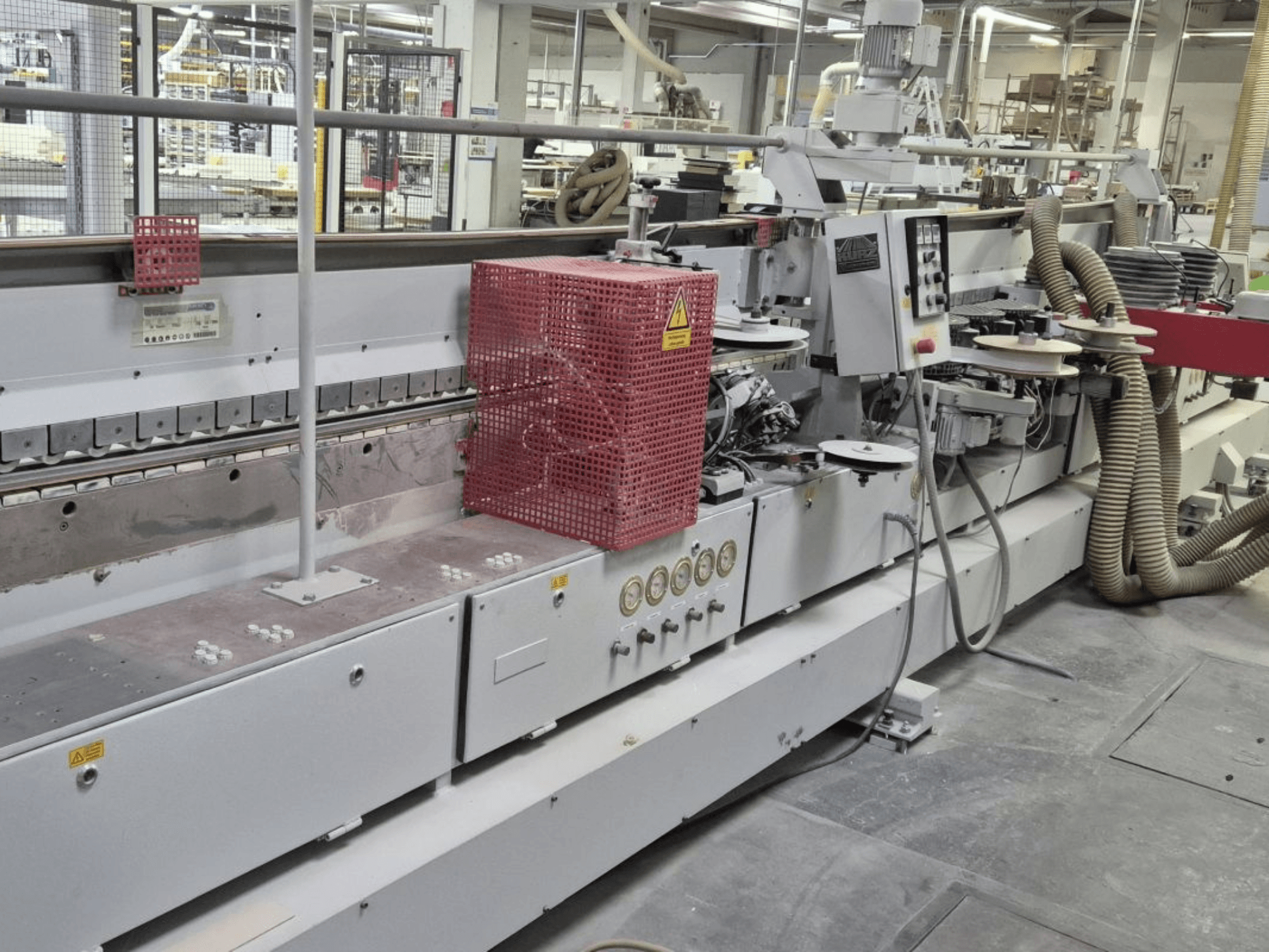 Máquina de aplicação de fita de borda com painel de controlo, mangueiras de sucção e uma cobertura de segurança vermelha, vista de um ângulo lateral.