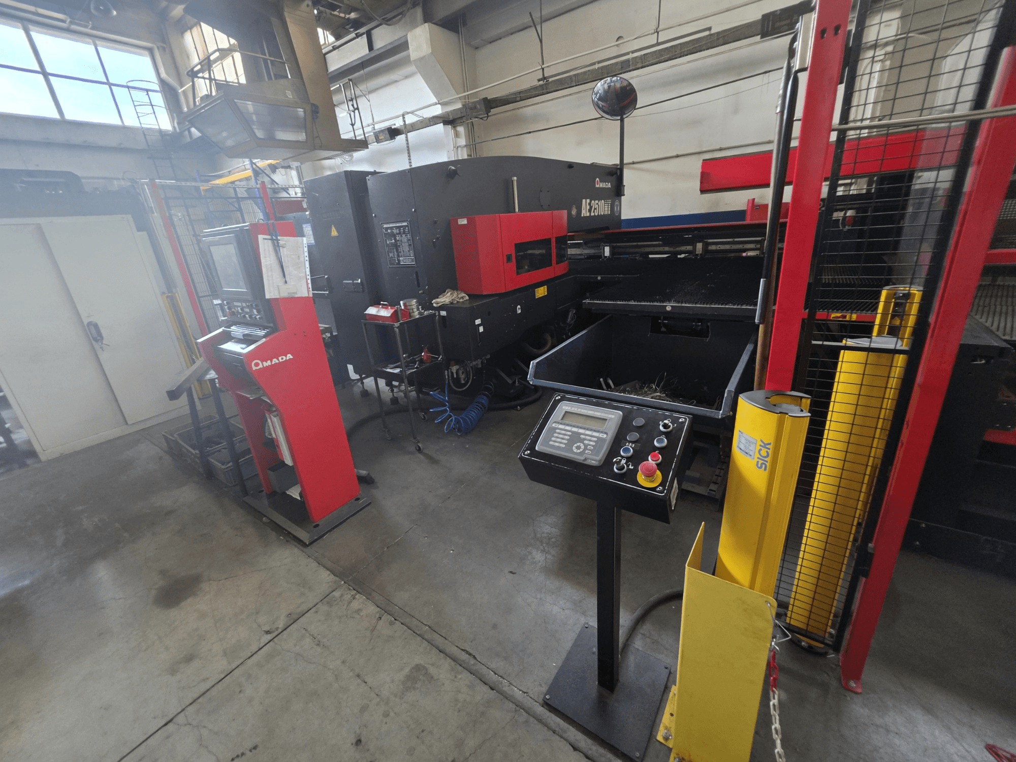 Máquina de perfuração CNC AMADA AE-2510NT, vista frontal com painel de controlo, dispositivos de segurança e ambiente industrial circundante.