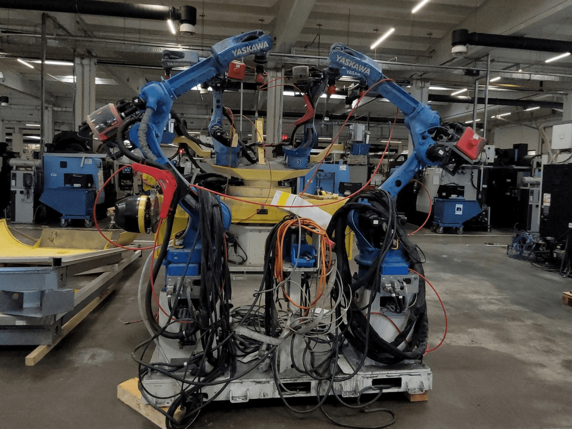 Célula de soldadura robótica Yaskawa RWC L1_OS 15330, vista frontal, com dois robôs azuis equipados com cabos extensos e ferramentas de soldadura.