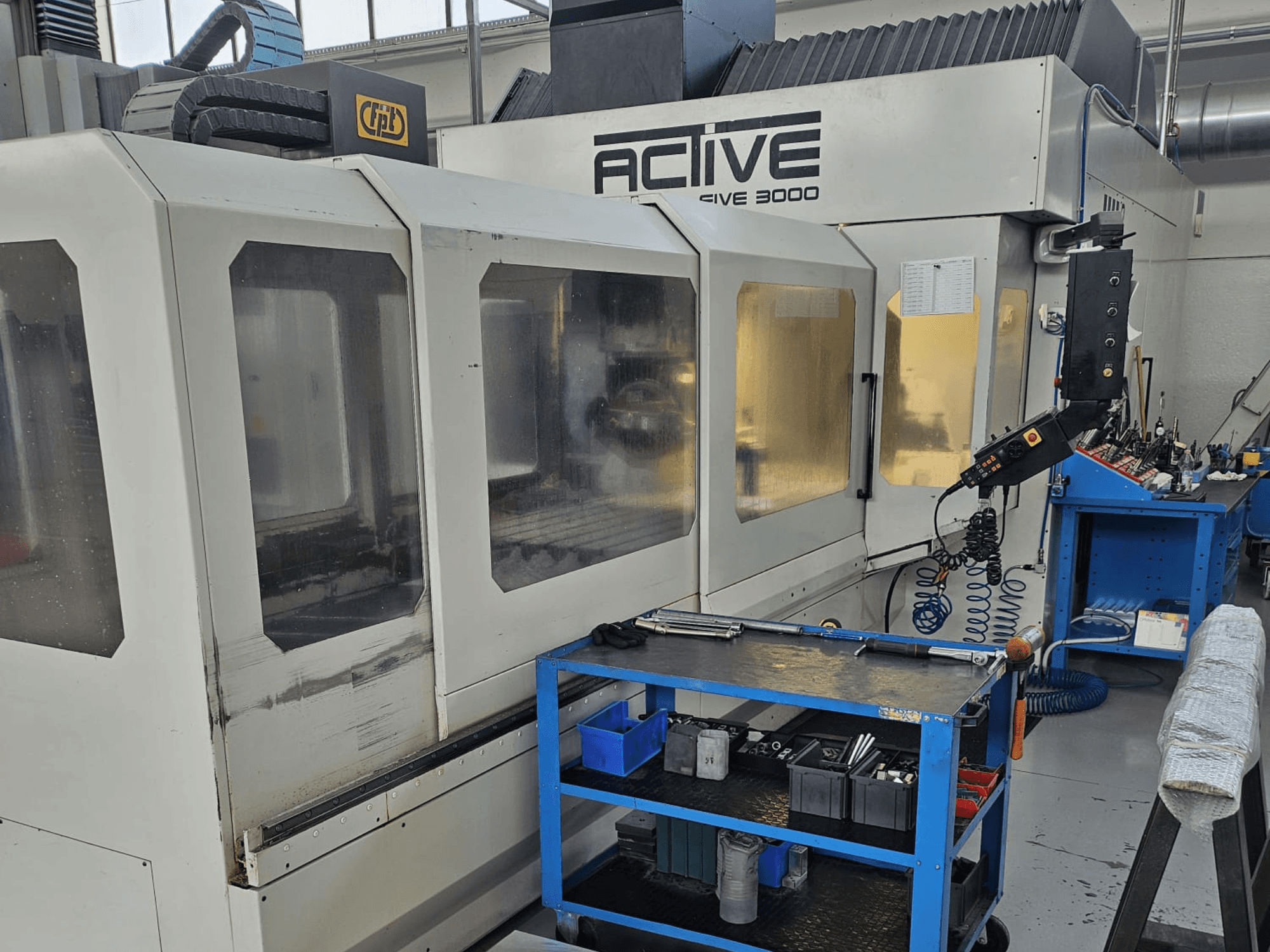 Máquina CNC Active 3000 numa oficina, vista frontal com ferramentas num carrinho azul, controlos visíveis e área de trabalho no interior.