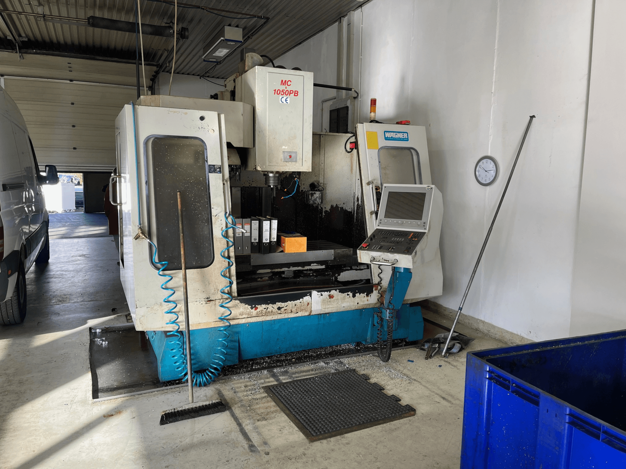 Máquina CNC WAGNER MC 1050PB na oficina, vista frontal, com painel de controlo e área de trabalho; mangueira de ar azul e ferramentas de limpeza visíveis.