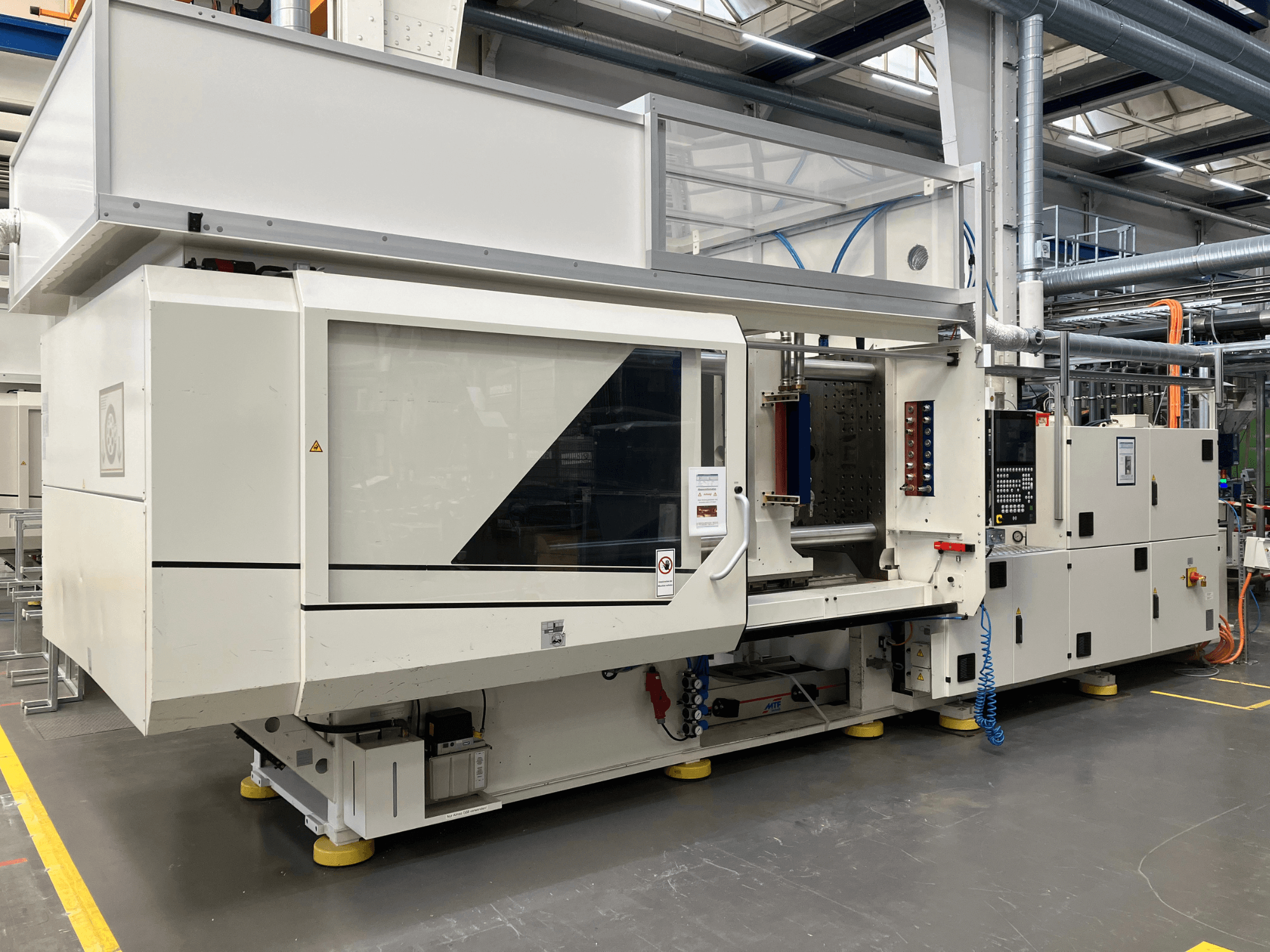 Vista lateral de uma máquina de moldagem por injeção, modelo AET, com painel de controlo e tampas transparentes, num ambiente industrial.