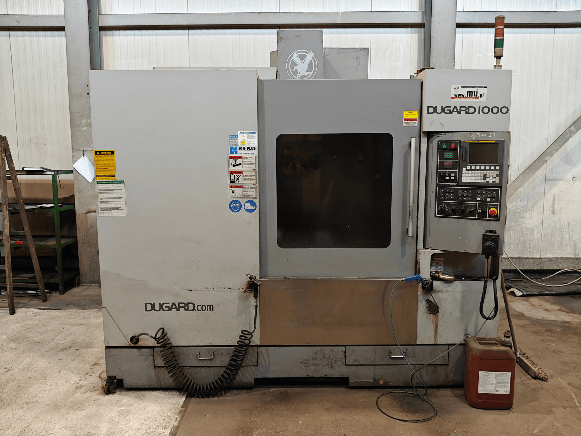 Máquina CNC Dugard 1000, vista frontal, mostrando o painel de controlo, etiquetas de segurança e recipiente de óleo, numa oficina.
