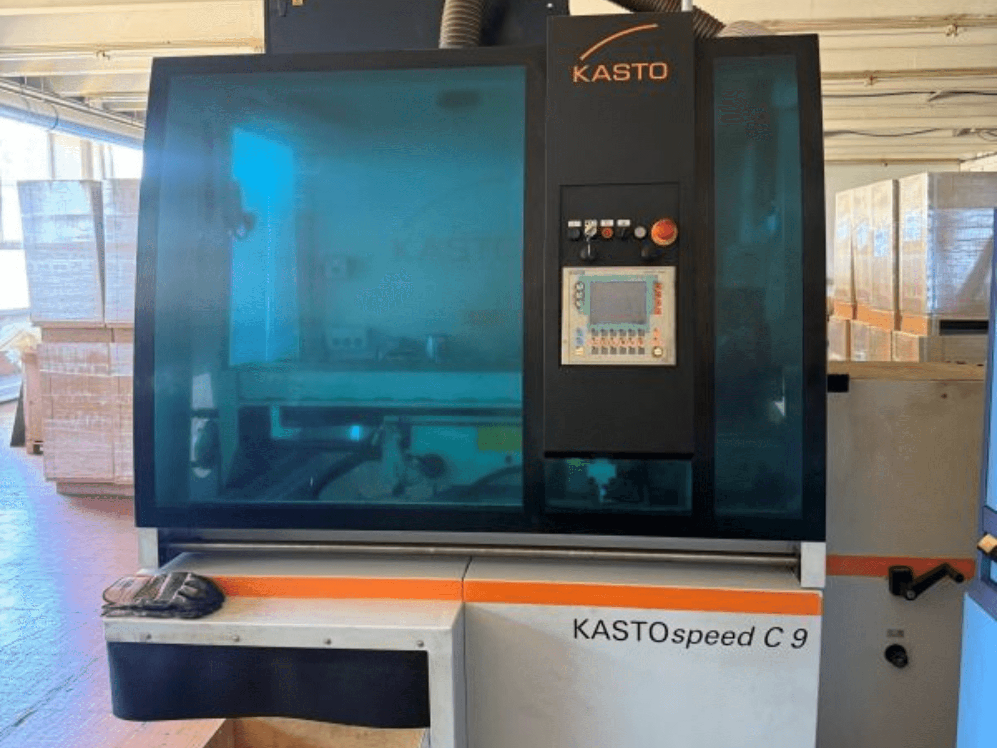 Vista frontal da serra circular para metal KASTO KASTOspeed C 9, com painel de controlo e vidro de segurança, tendo como pano de fundo caixas de madeira.