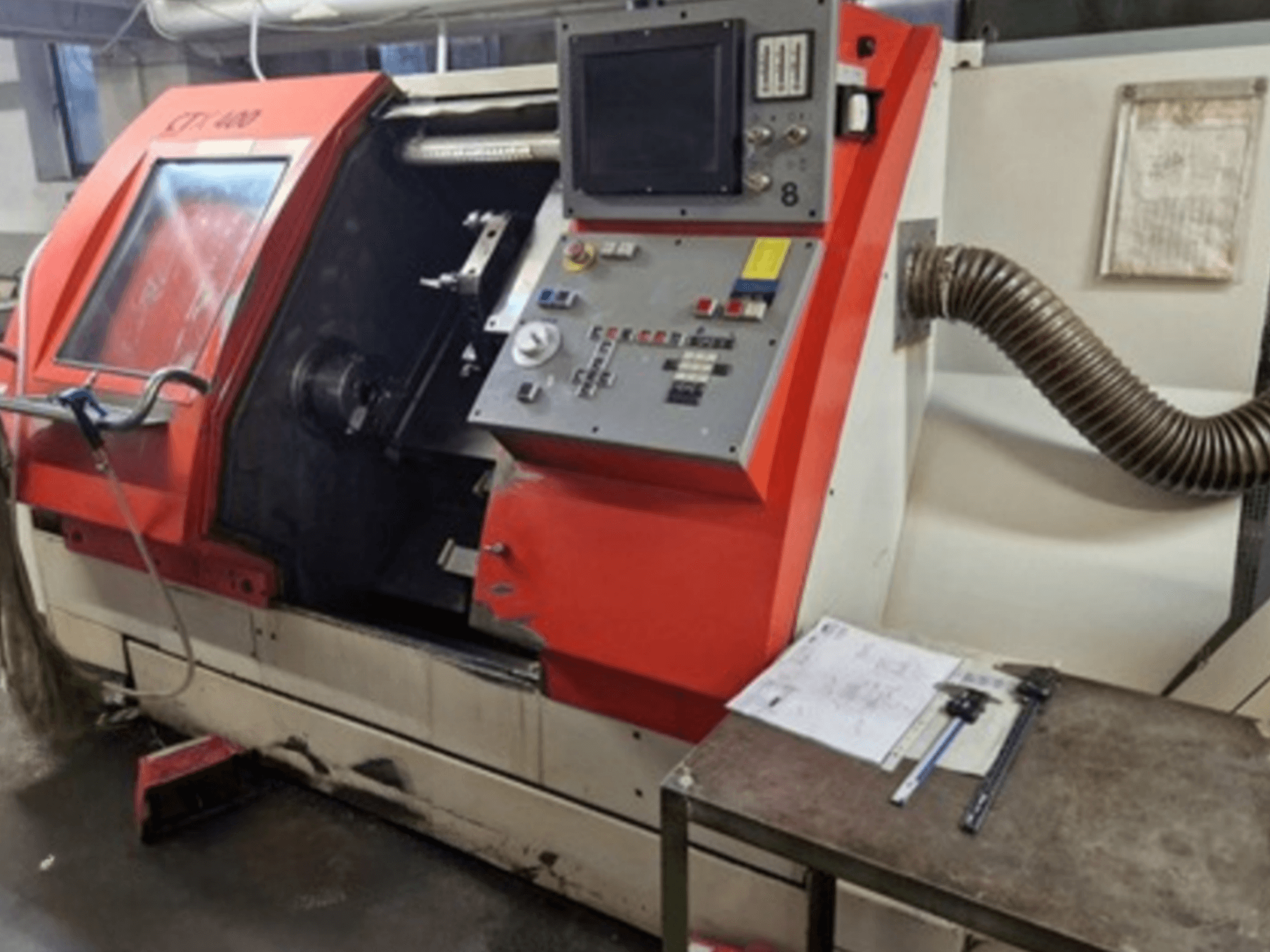 Torneiro horizontal Gildemeister CTX 400, vista frontal com painel de controlo e tubo de escape. Inclui ferramentas de medição na mesa.