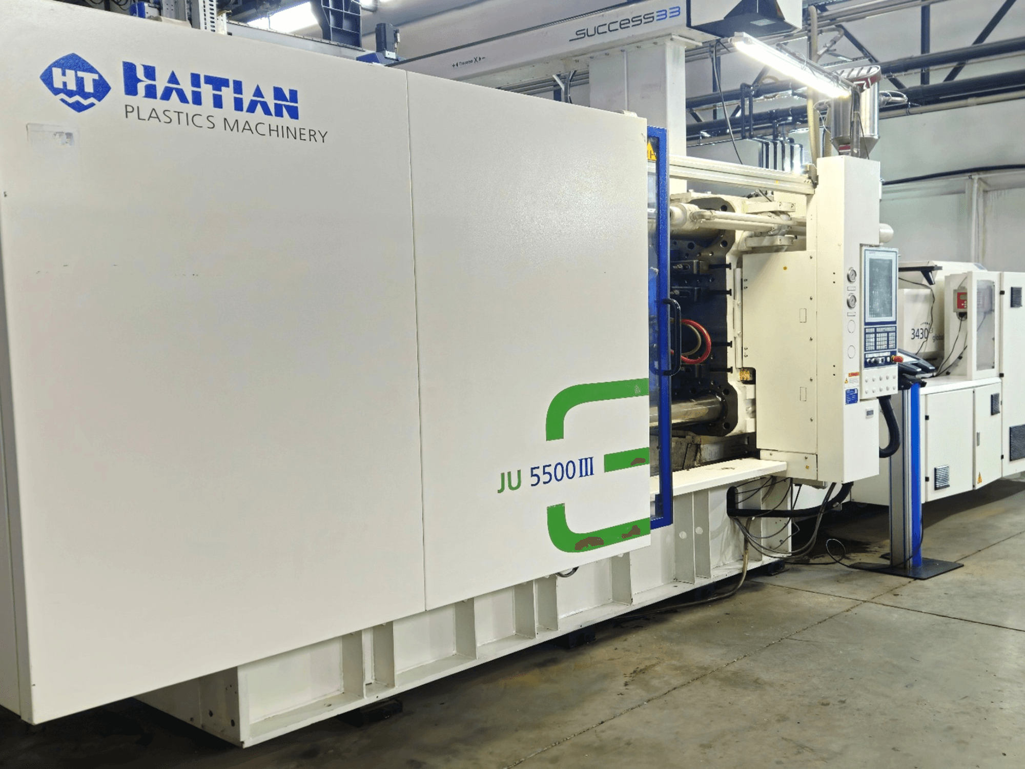 Máquina de plástico JU 5500III da Haitian numa fábrica, vista frontal mostrando o painel de controlo e a área de produção.